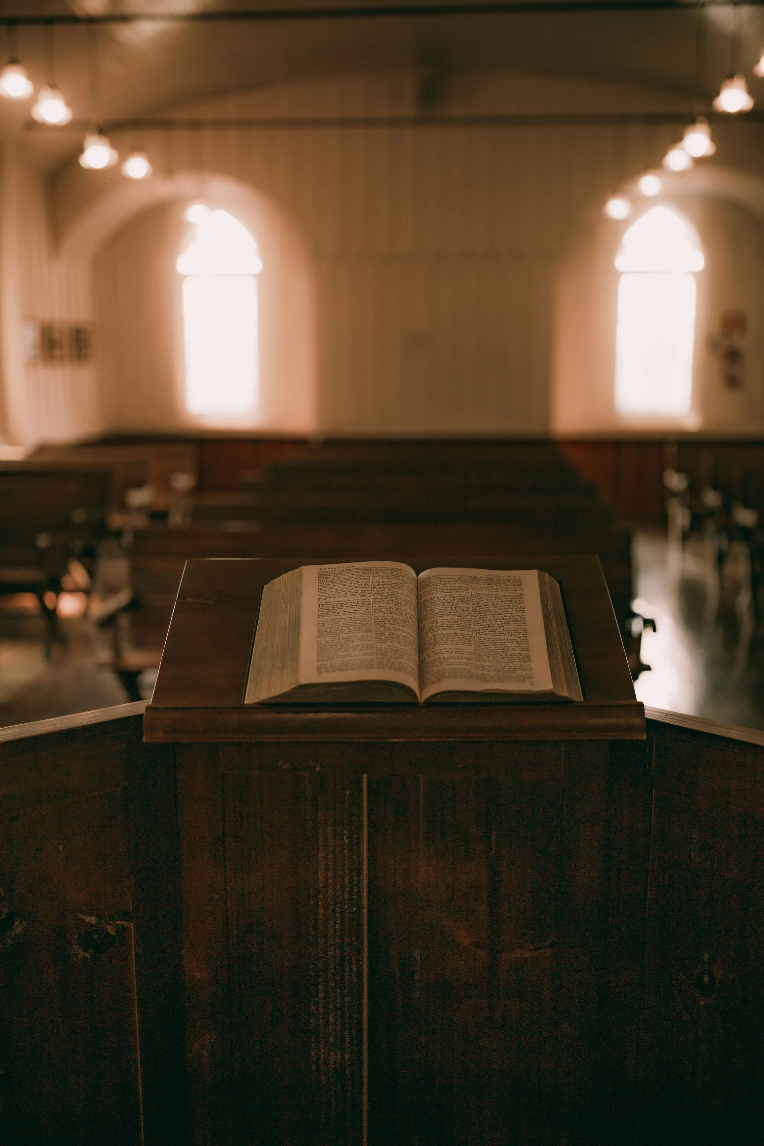 An open Bible on a wooden lectern in a church or chapel, with rows of wooden pews and tall, arched stained glass windows in the background, softly illuminated by warm light.