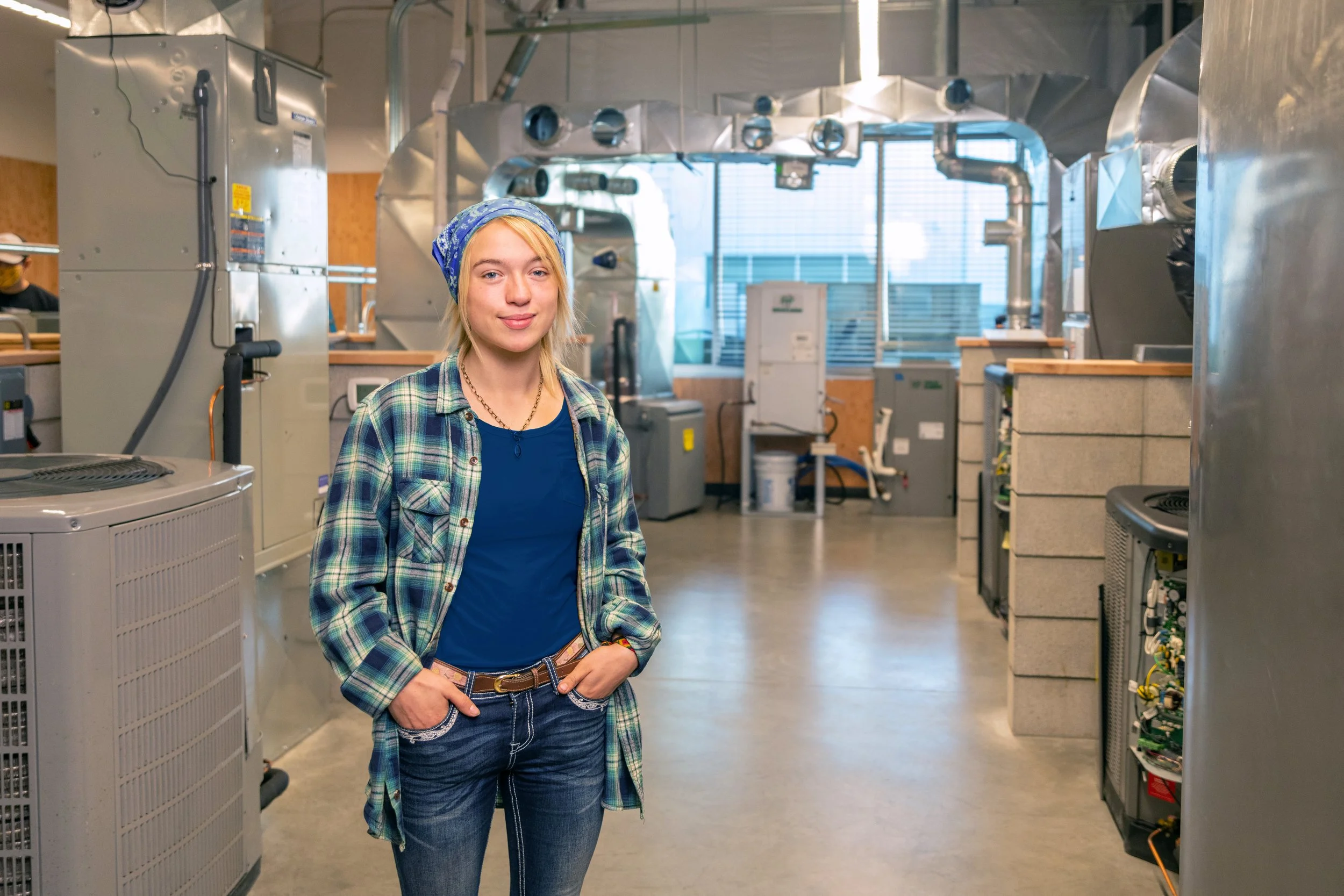 A young woman with blonde hair, wearing a blue bandana, blue shirt, and plaid shirt, standing with her hands in her pockets inside an industrial or commercial laundry room or similar facility.