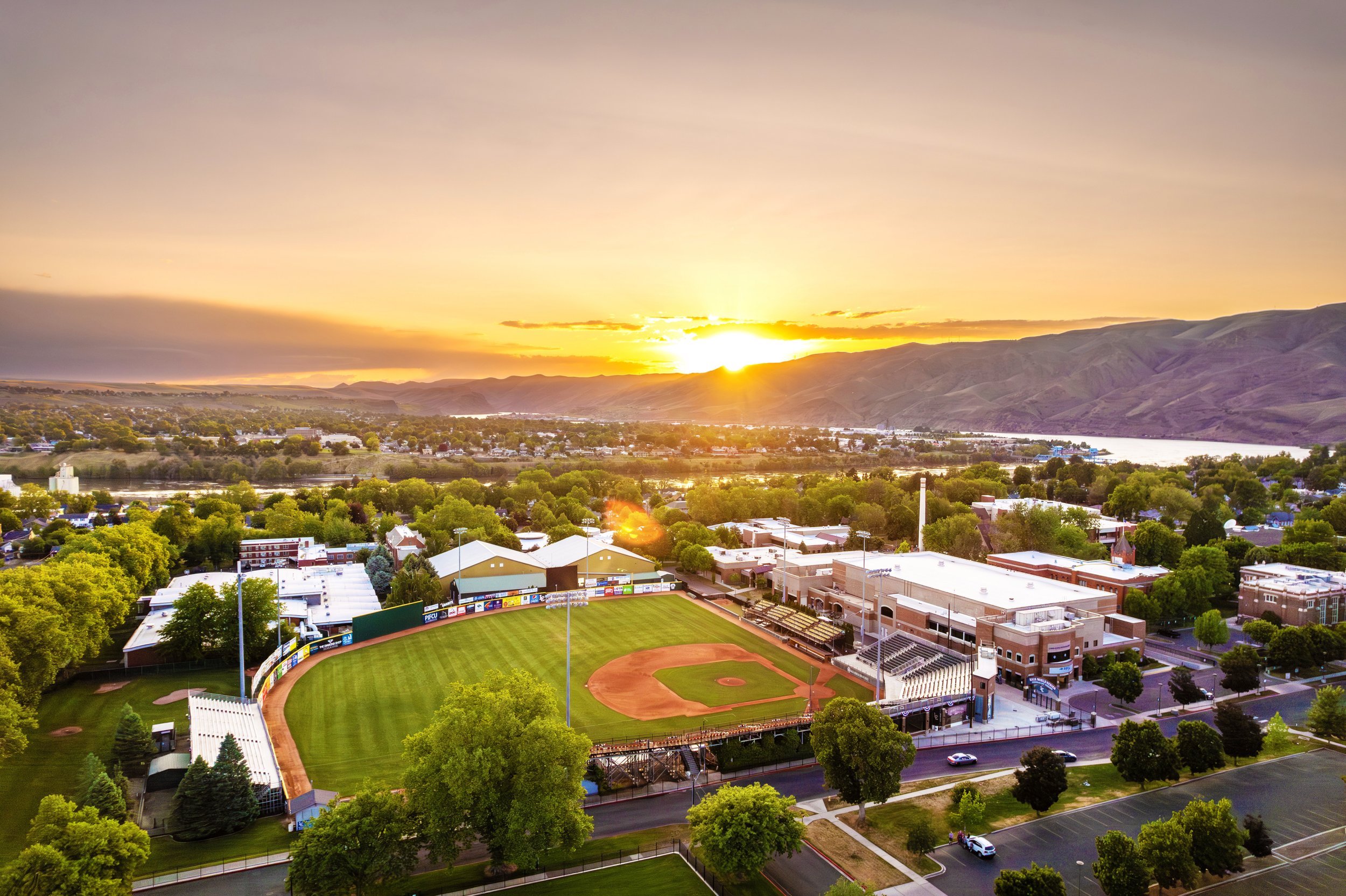 Aerial view of a baseball stadium at sunset, surrounded by trees and city buildings, with mountains and a river in the background.