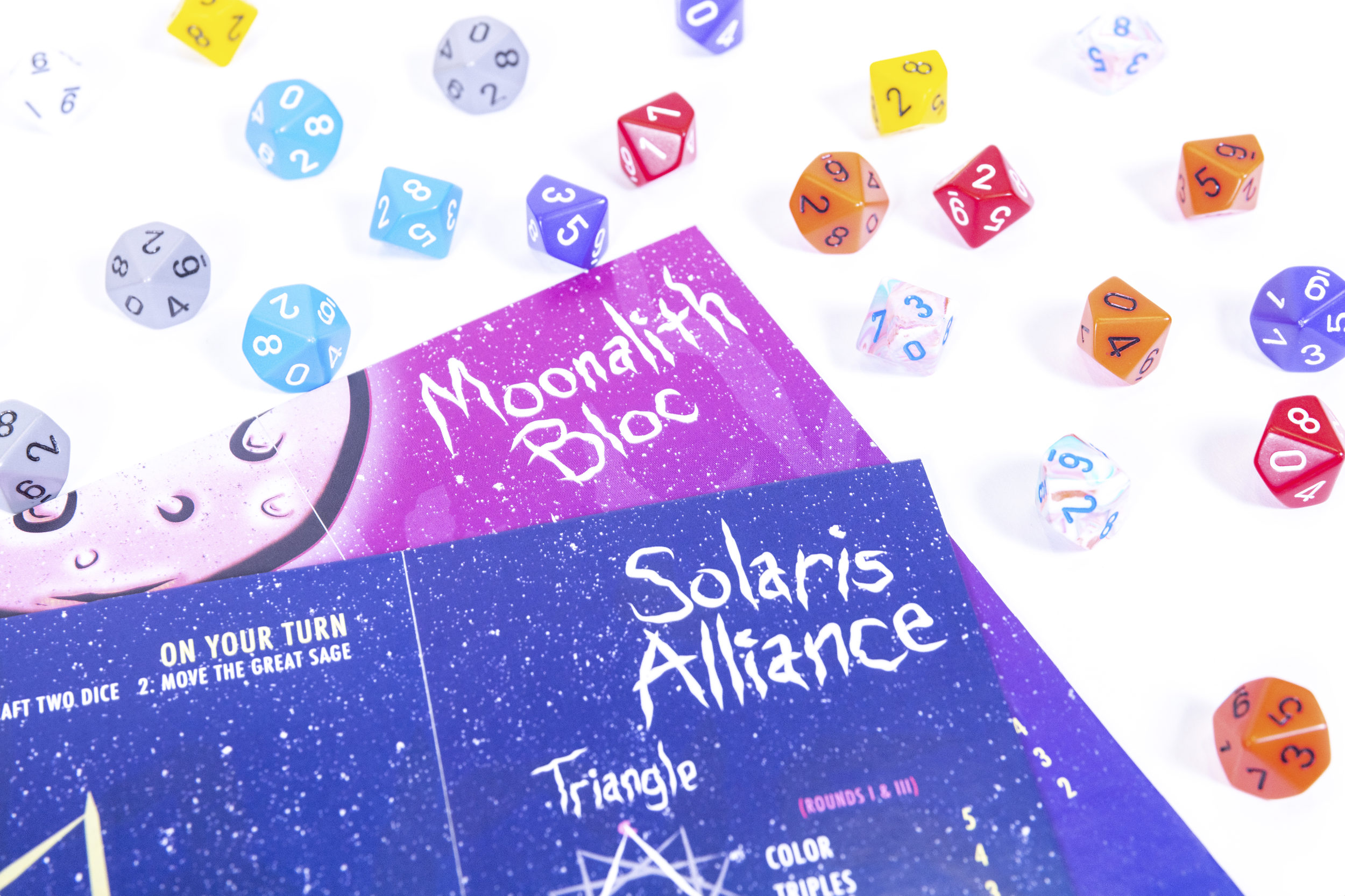 Colorful tabletop game components including multicolored polyhedral dice and a game instruction sheet titled 'Moonlitith Bloc' and 'Solaris Alliance' on a white background.