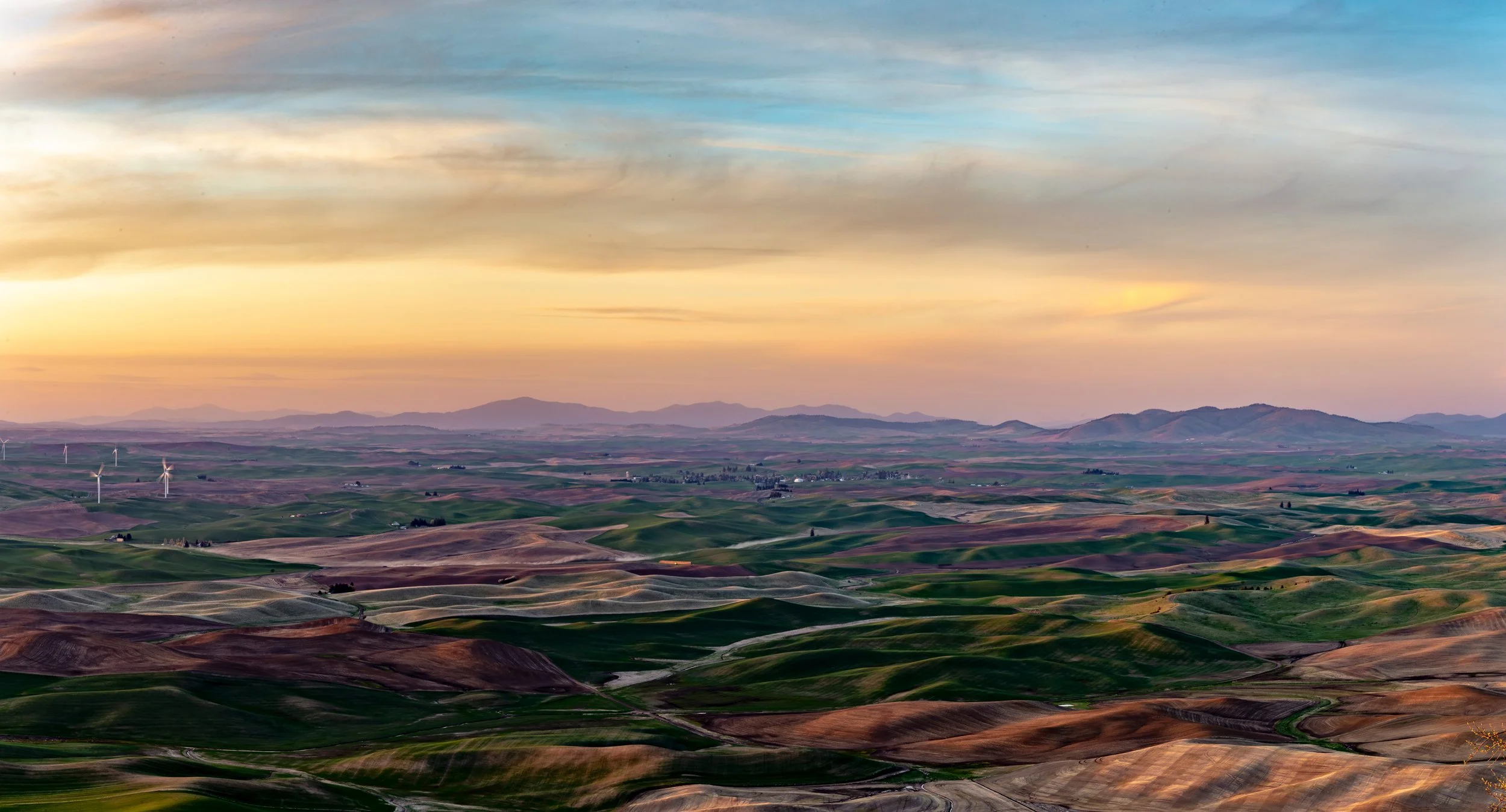A landscape view of rolling hills at sunset with wind turbines in the distance and a partly cloudy sky.