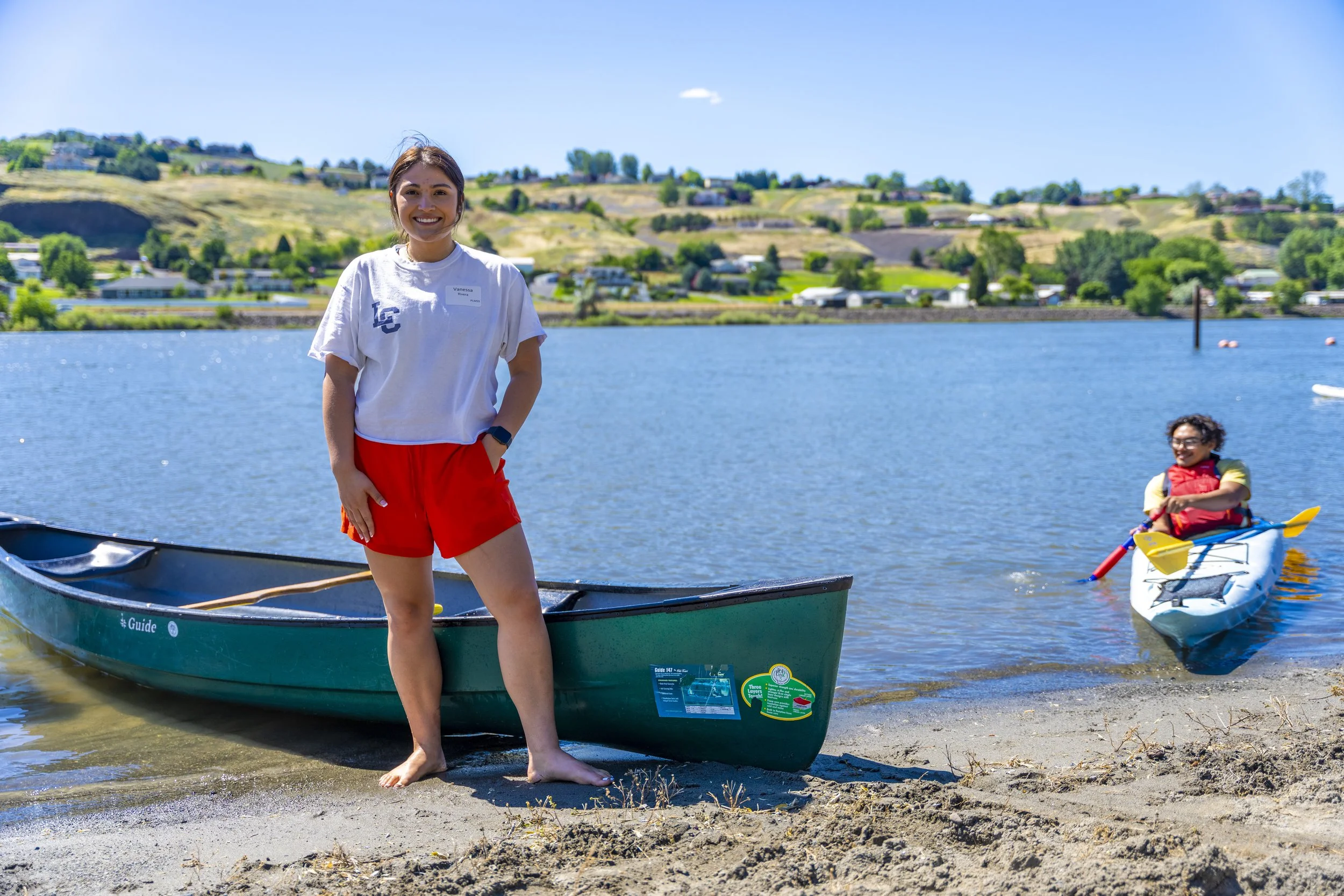 A woman with brown hair smiling stands barefoot on a sandy shore beside a green canoe, with a young girl paddling a blue and white kayak in the water behind her, on a sunny day with a hilly landscape in the background.