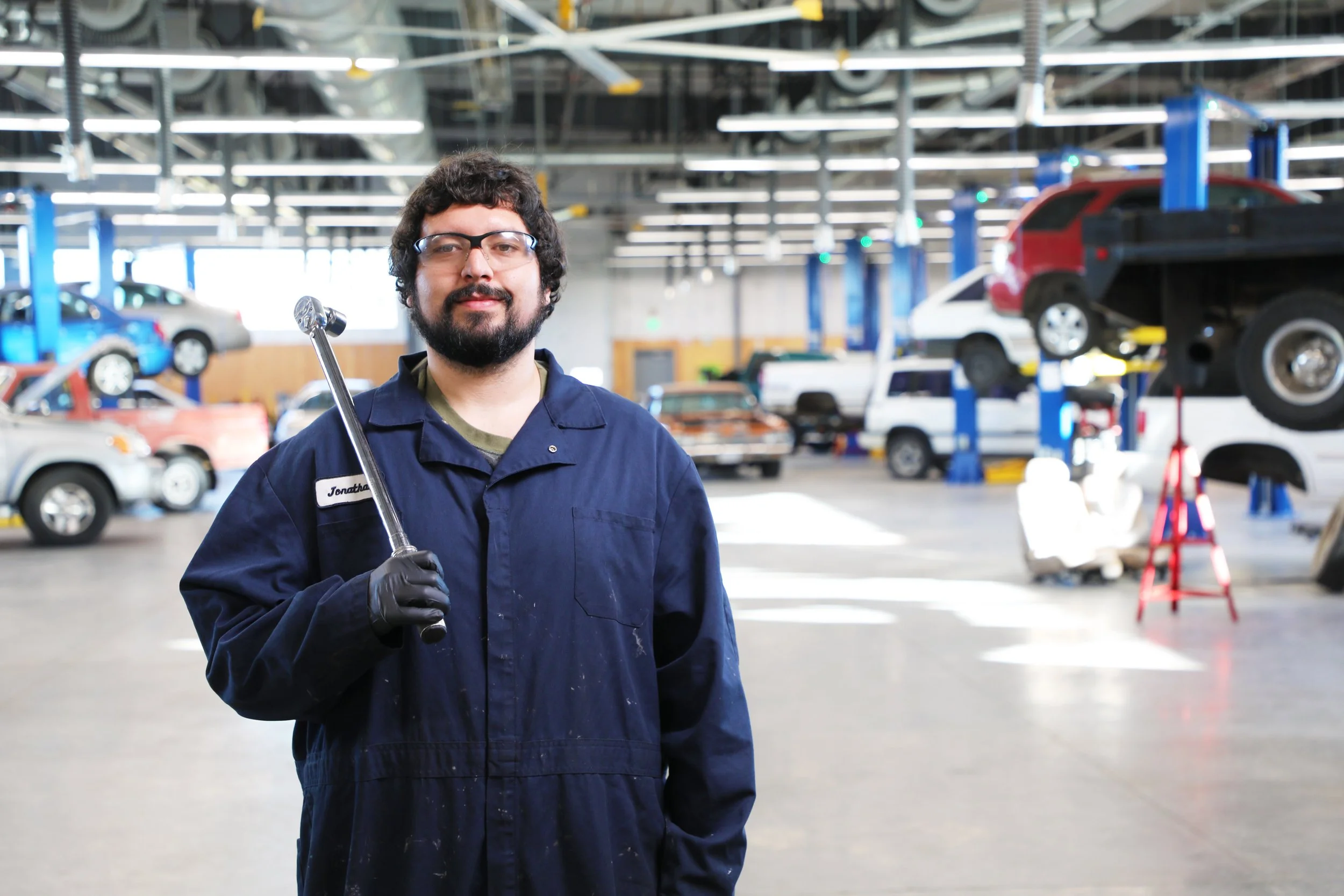 An auto mechanic with glasses and a beard holding a wrench in a car repair shop with cars lifted on hydraulic lifts.