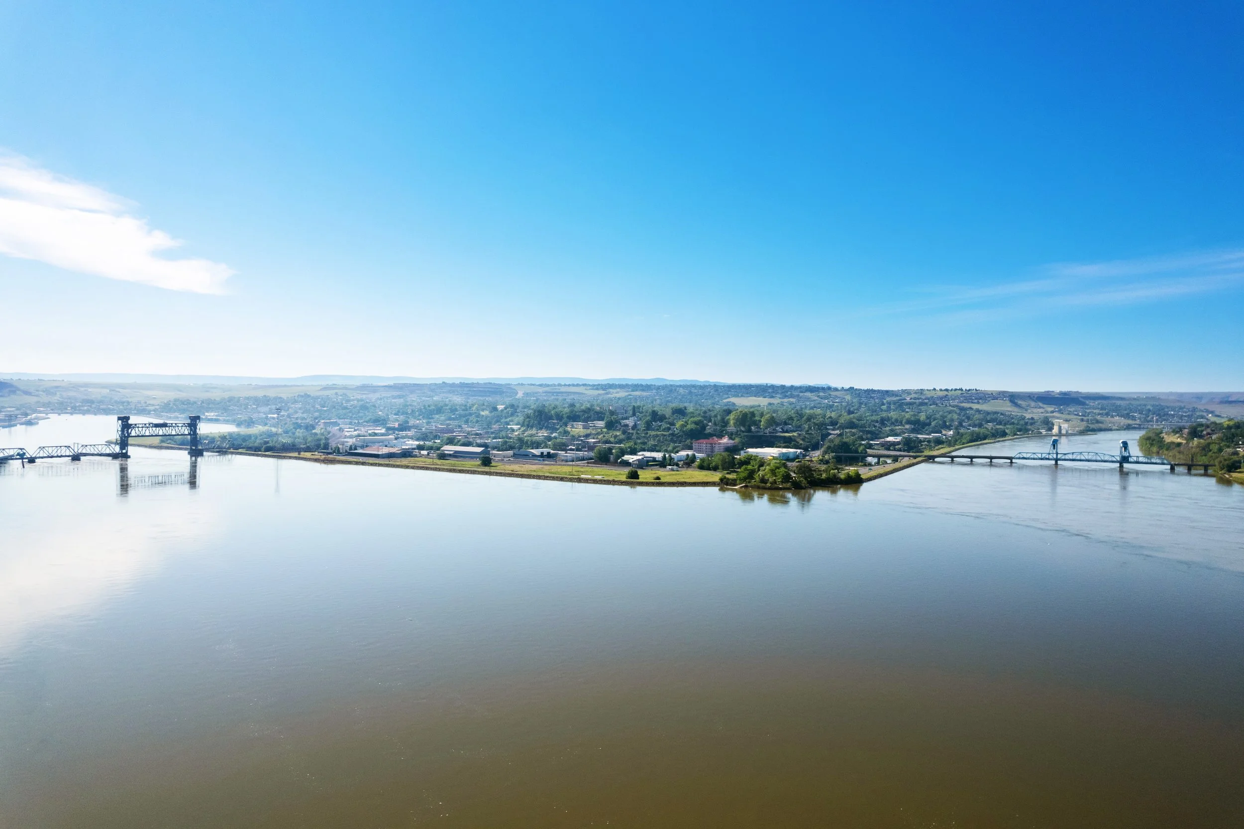 A wide river with a distant view of the city skyline, including bridges and industrial buildings, under a bright blue sky.