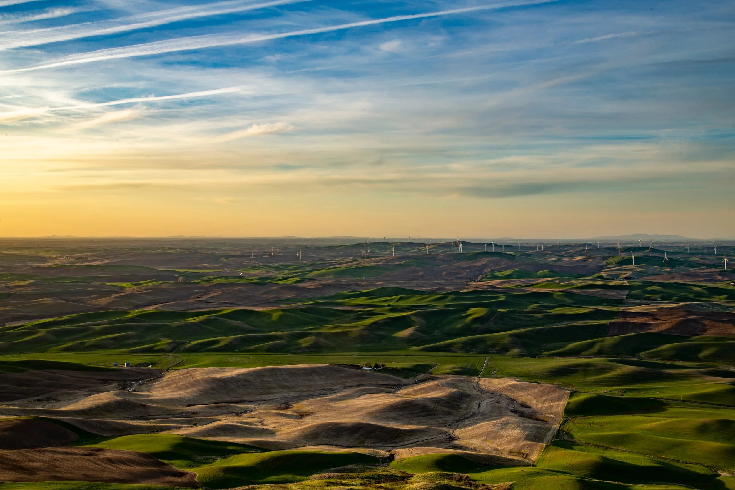Sunset over rolling green and brown hills with wind turbines in the distance under a partly cloudy sky.