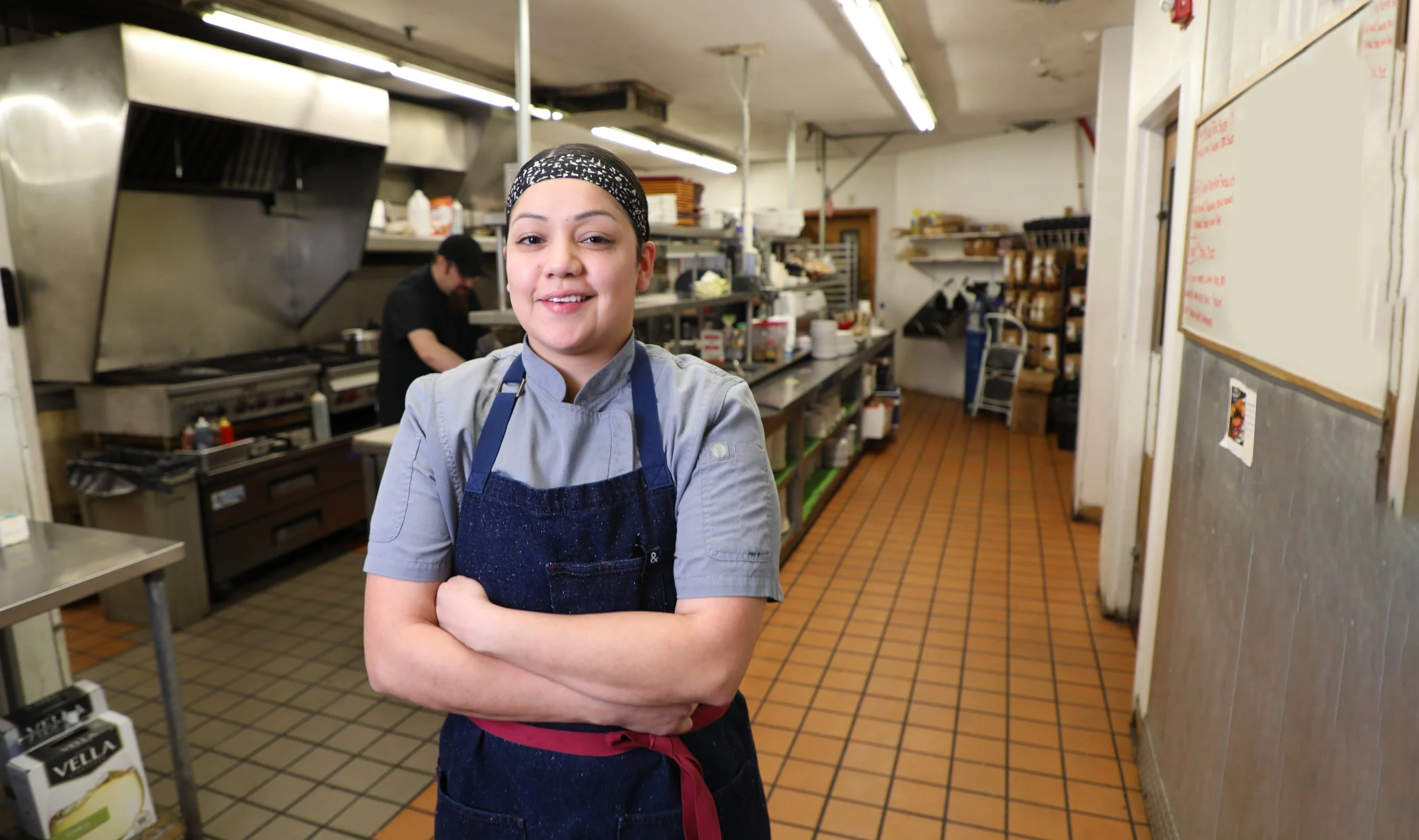 Smiling woman in a restaurant kitchen wearing a gray shirt, blue apron, and hair cover, with kitchen equipment and a chef in the background.