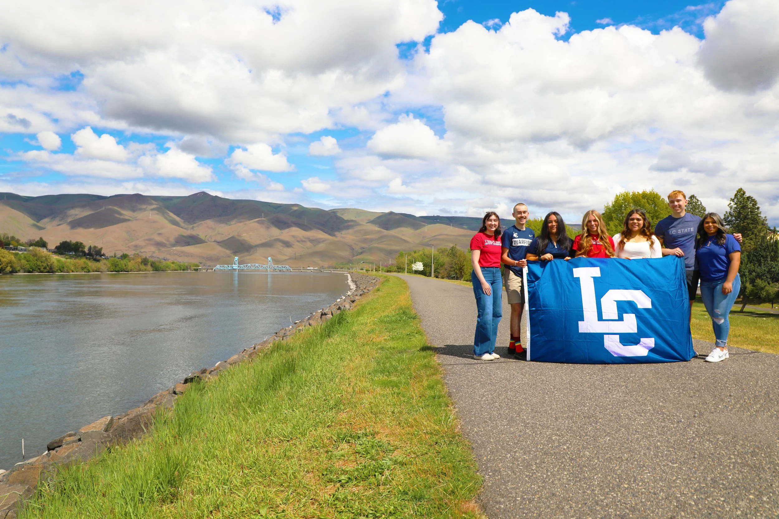 A group of six young people standing on a paved path beside a river, holding a blue LSU flag and smiling at the camera, with hills and a bridge in the background on a partly cloudy day.