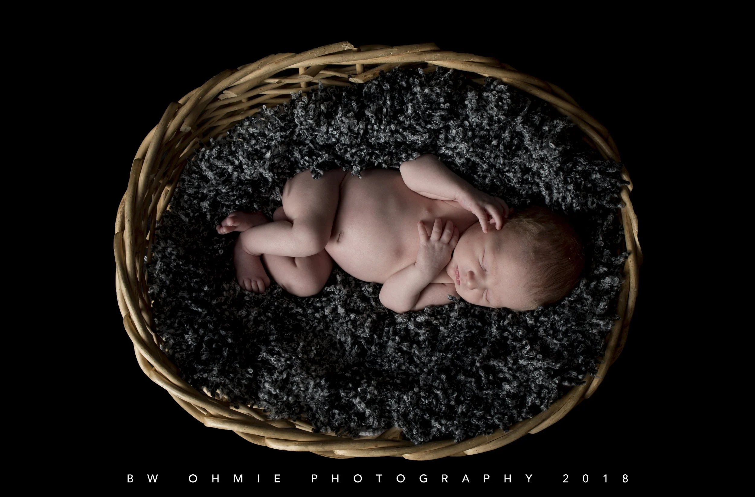 A newborn baby sleeping in a wicker basket on a soft black blanket with white speckles.