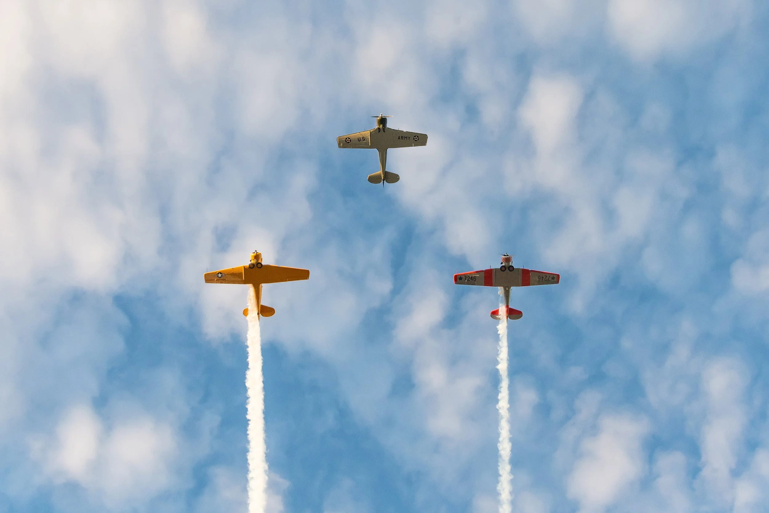 Three small planes flying in formation, leaving white smoke trails in a blue sky with scattered clouds.