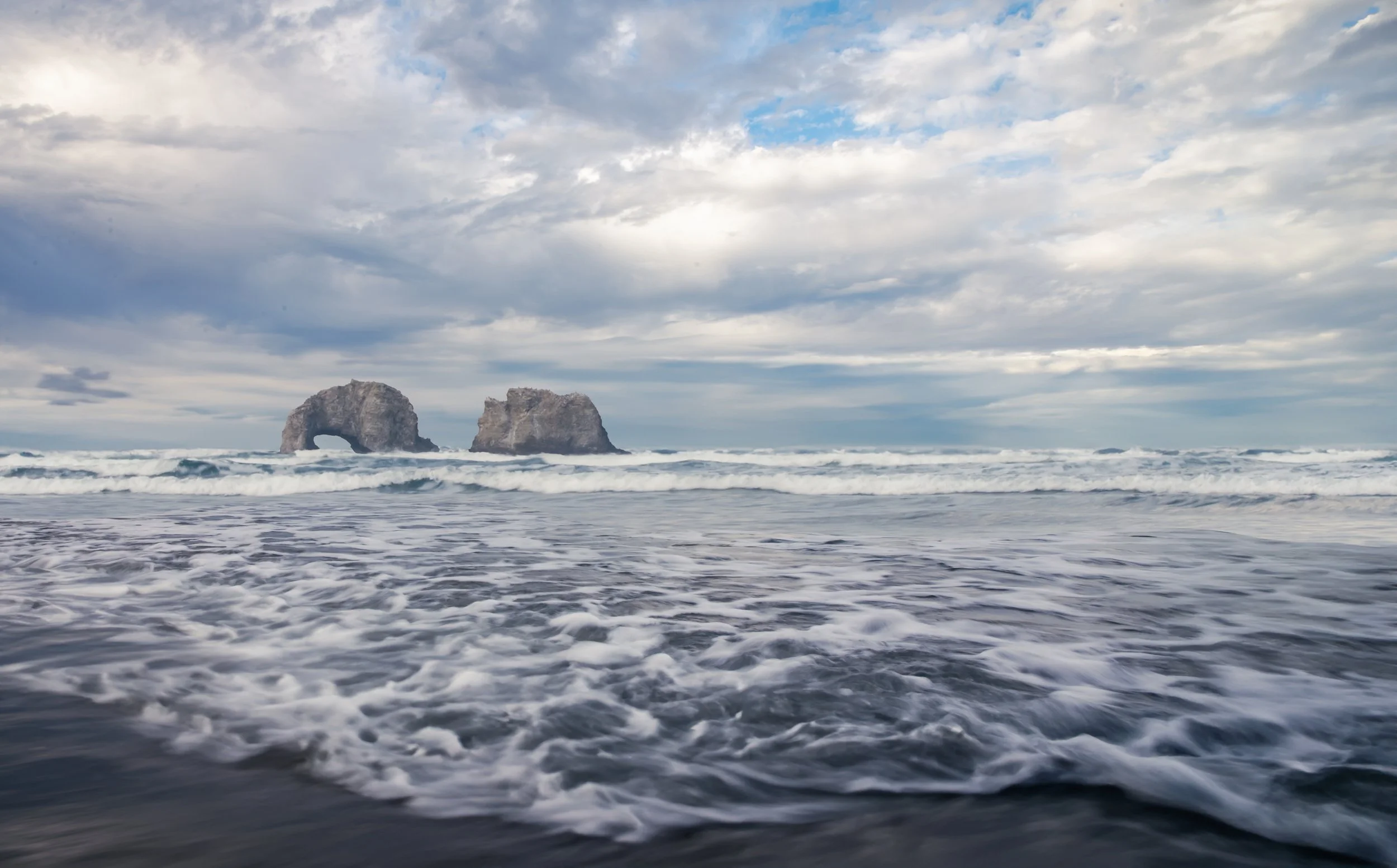 Ocean with two large rock formations in the distance, waves crashing on the shore, and a cloudy sky.