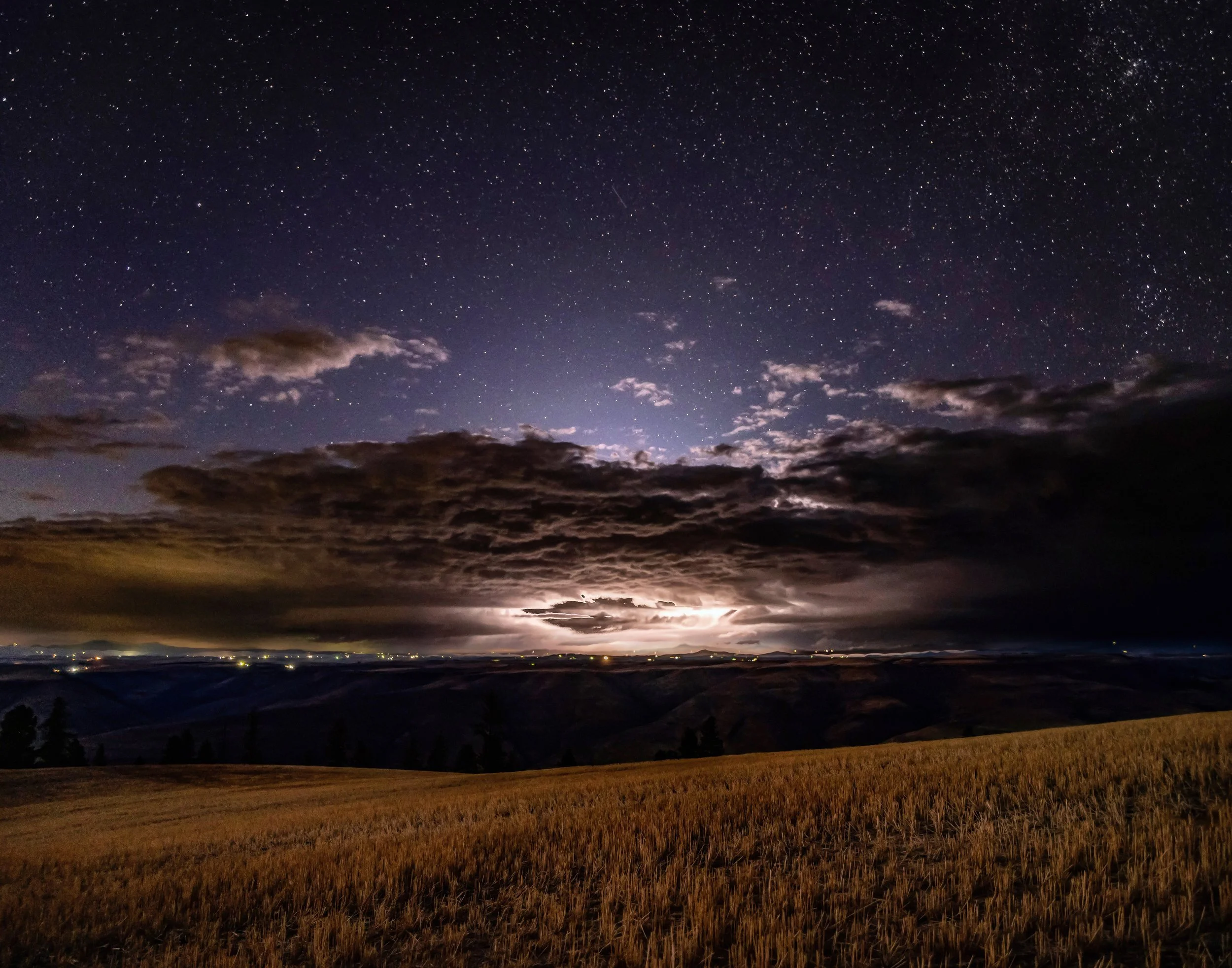 Night sky filled with stars over open fields and distant mountains, with clouds illuminated by lightning.
