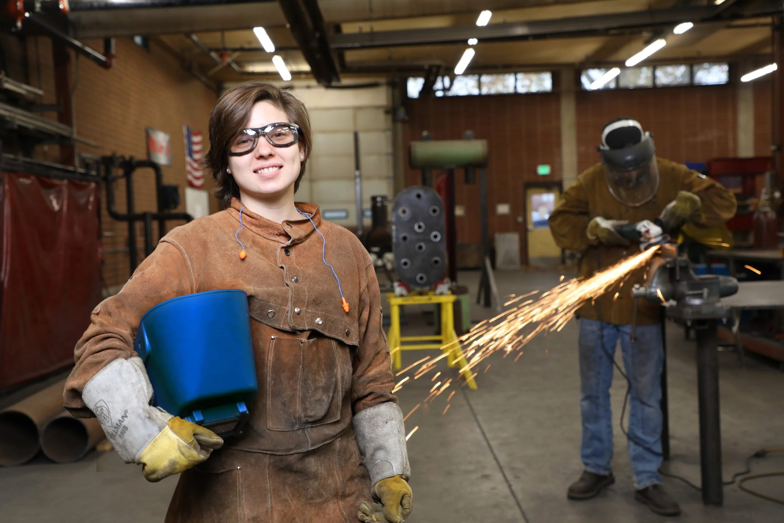 A woman and a man in a workshop, the woman wearing protective gloves, glasses, and holding a welding shield, while the man is welding with sparks flying.