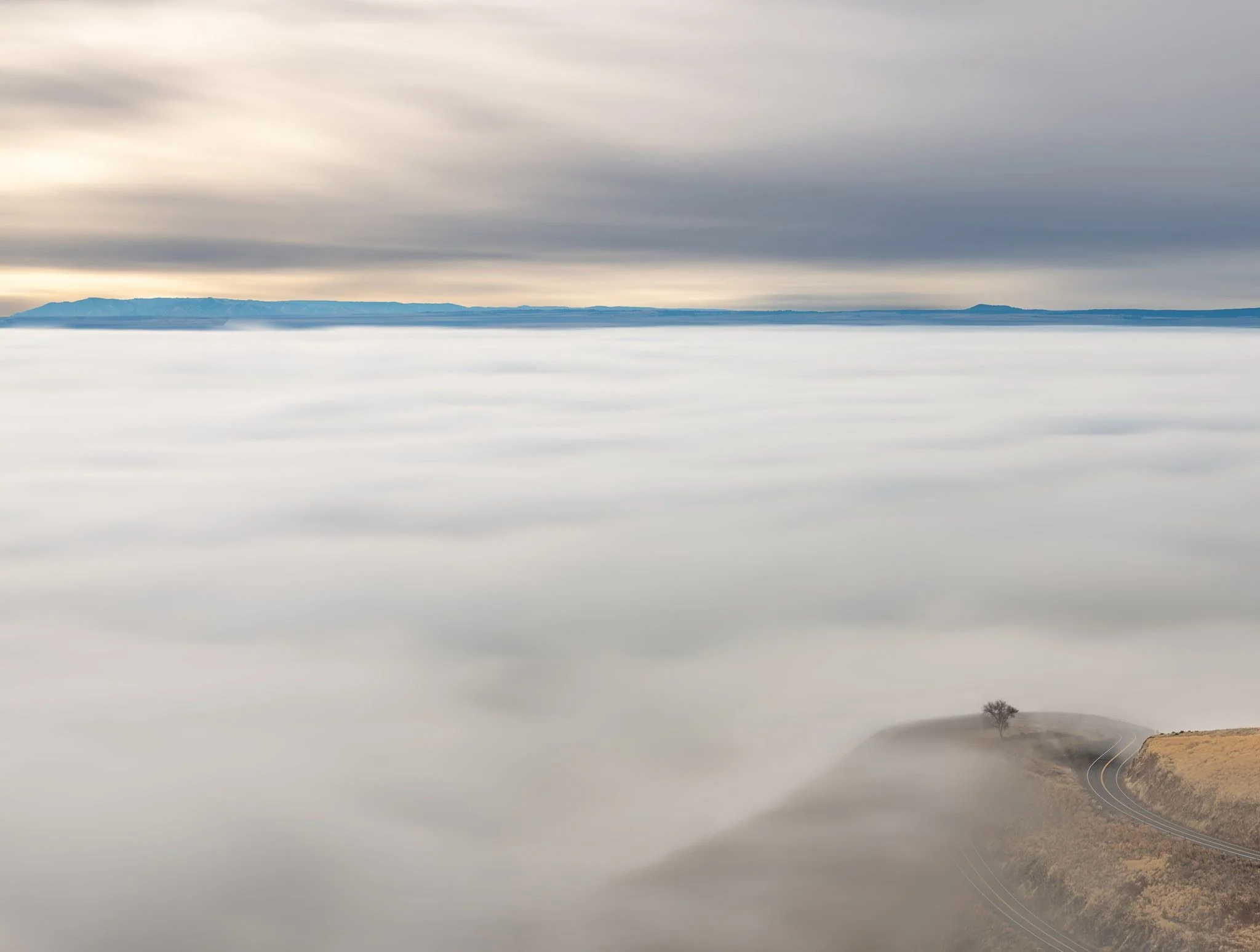 A vast landscape with fog covering the ground, a body of water in the background, mountains under a cloudy sky, and a small hill with a lone tree and a curved road on the lower right.