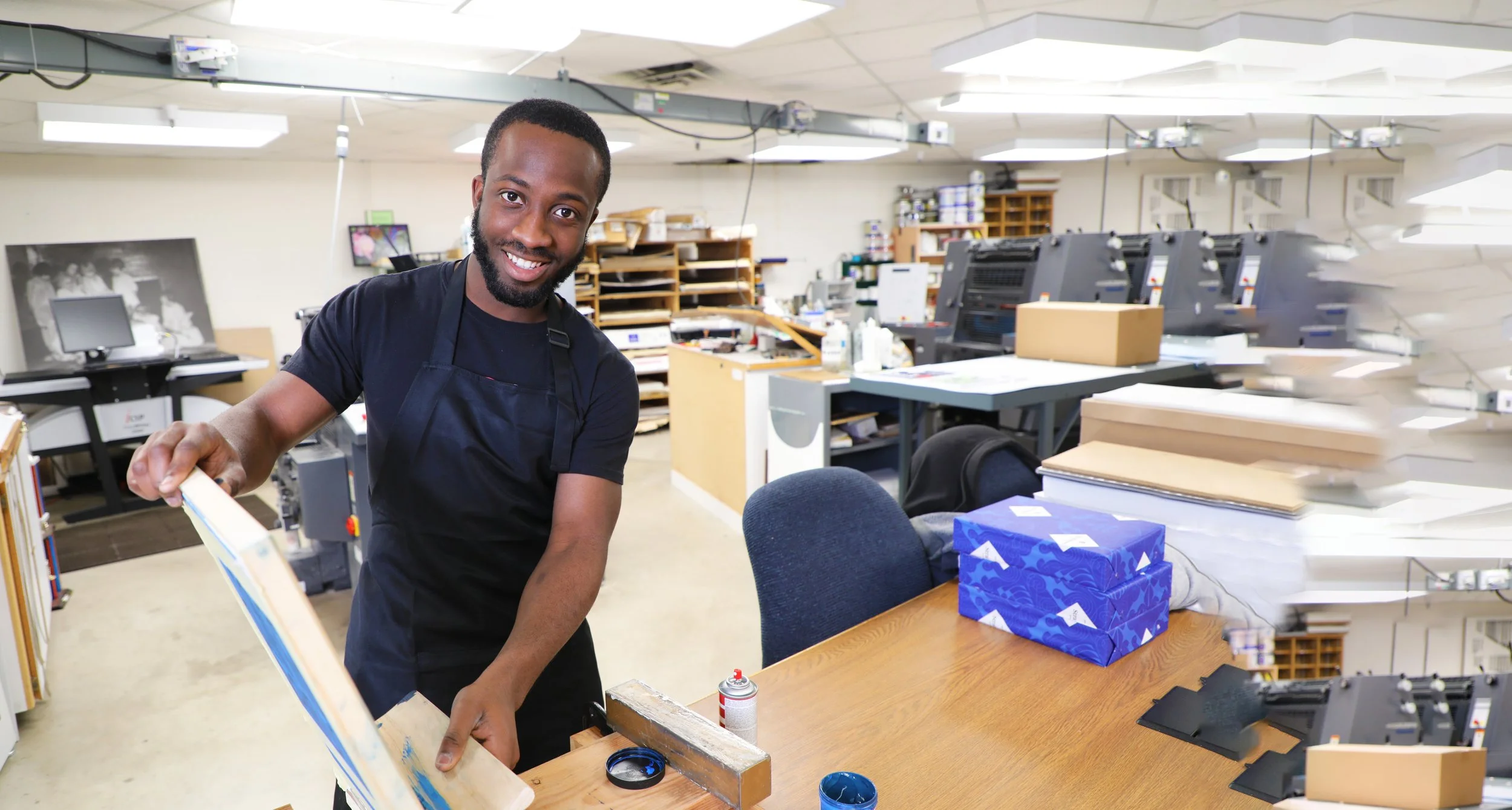 A smiling man in a black shirt and apron working at a printing or packaging workshop, surrounded by printing machines, shelves with supplies, and packages on a wooden table.