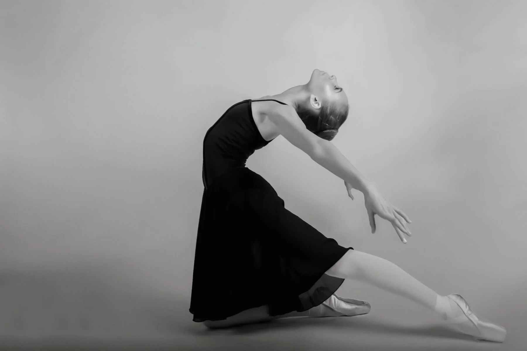 A female ballet dancer in a black leotard and skirt performing a pose, with her head tilted back, arms extended, and one leg stretched out on the floor, captured in a side profile. The image is in black and white.