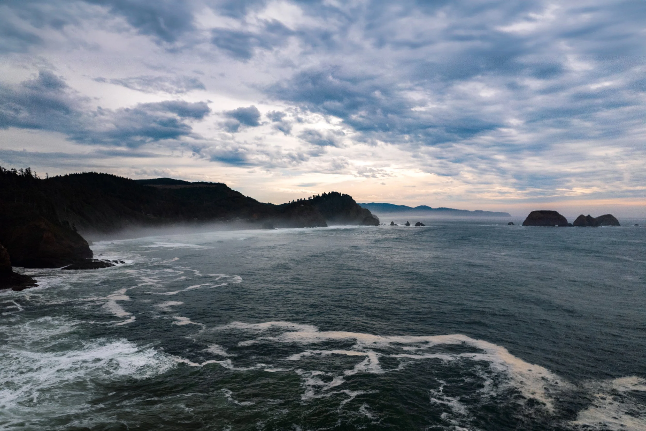 Coastal view with rugged cliffs, rocky islands, and ocean waves under a cloudy sky.