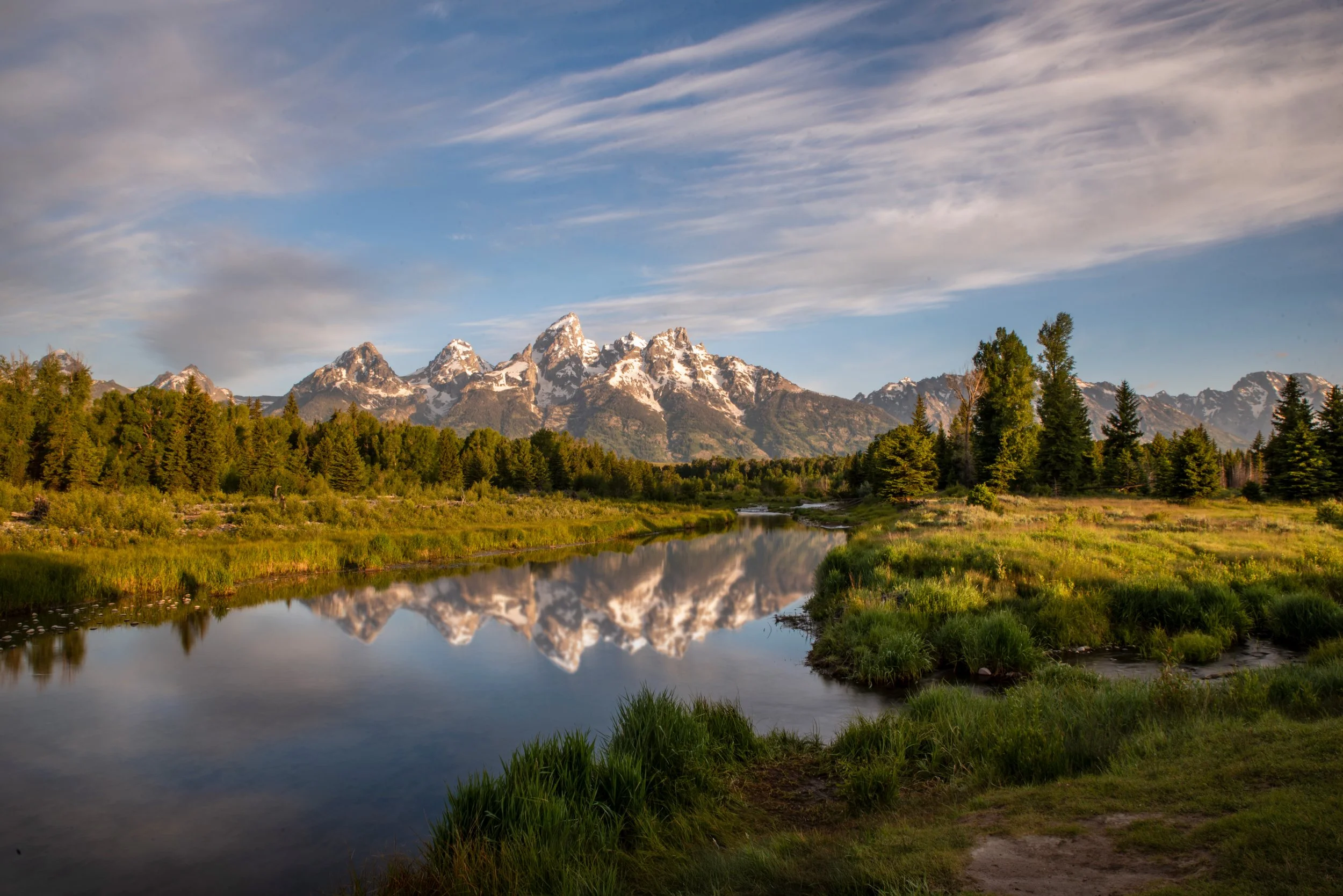 Snow-capped mountains reflected in a calm river surrounded by green trees and grass in a valley.