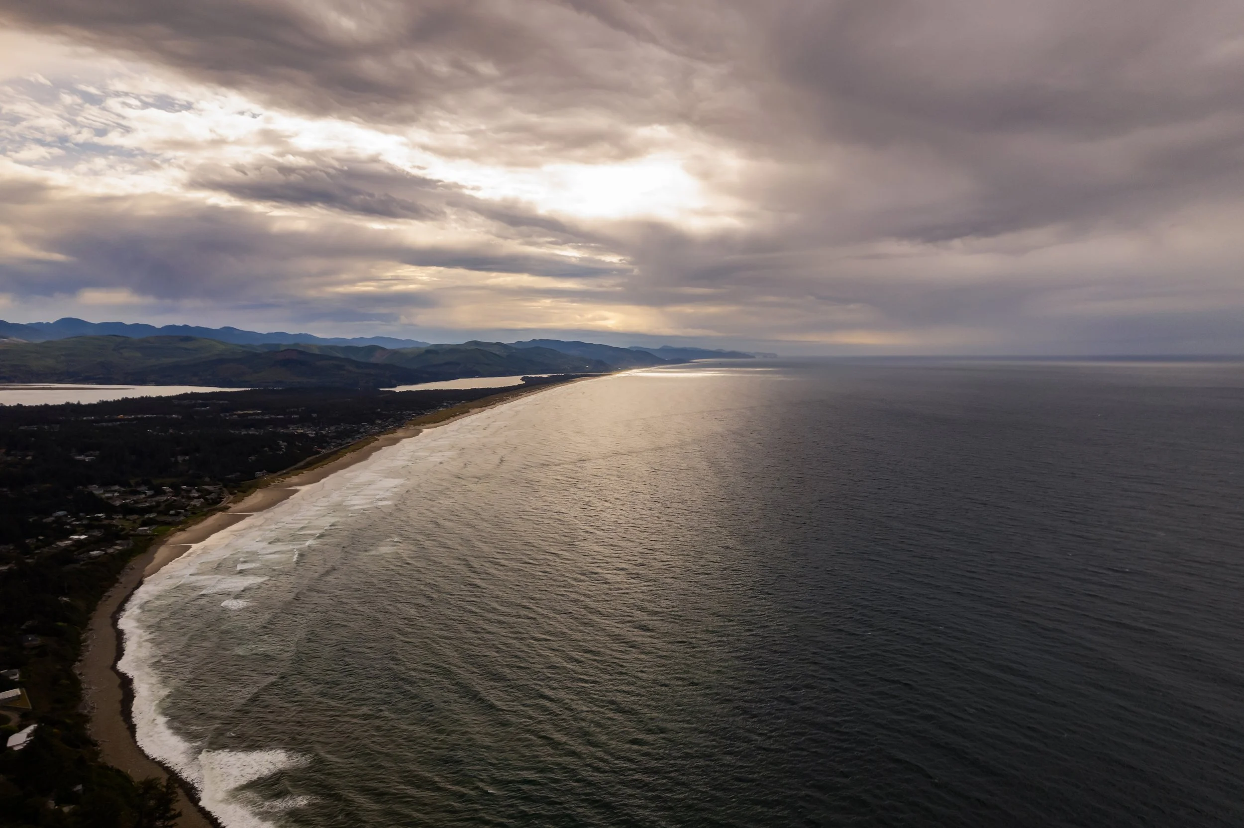 Aerial view of a beach with waves, a distant shoreline with a town and mountains under a cloudy sky.