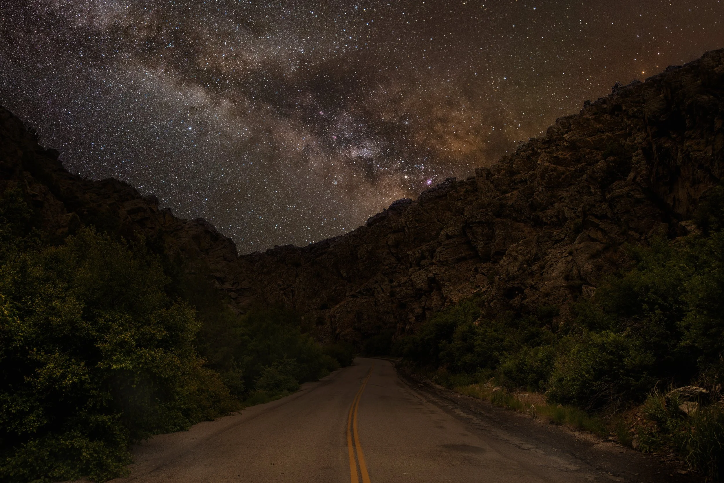 A scenic night view of a winding road through rocky mountains with a starry sky and the Milky Way galaxy overhead.