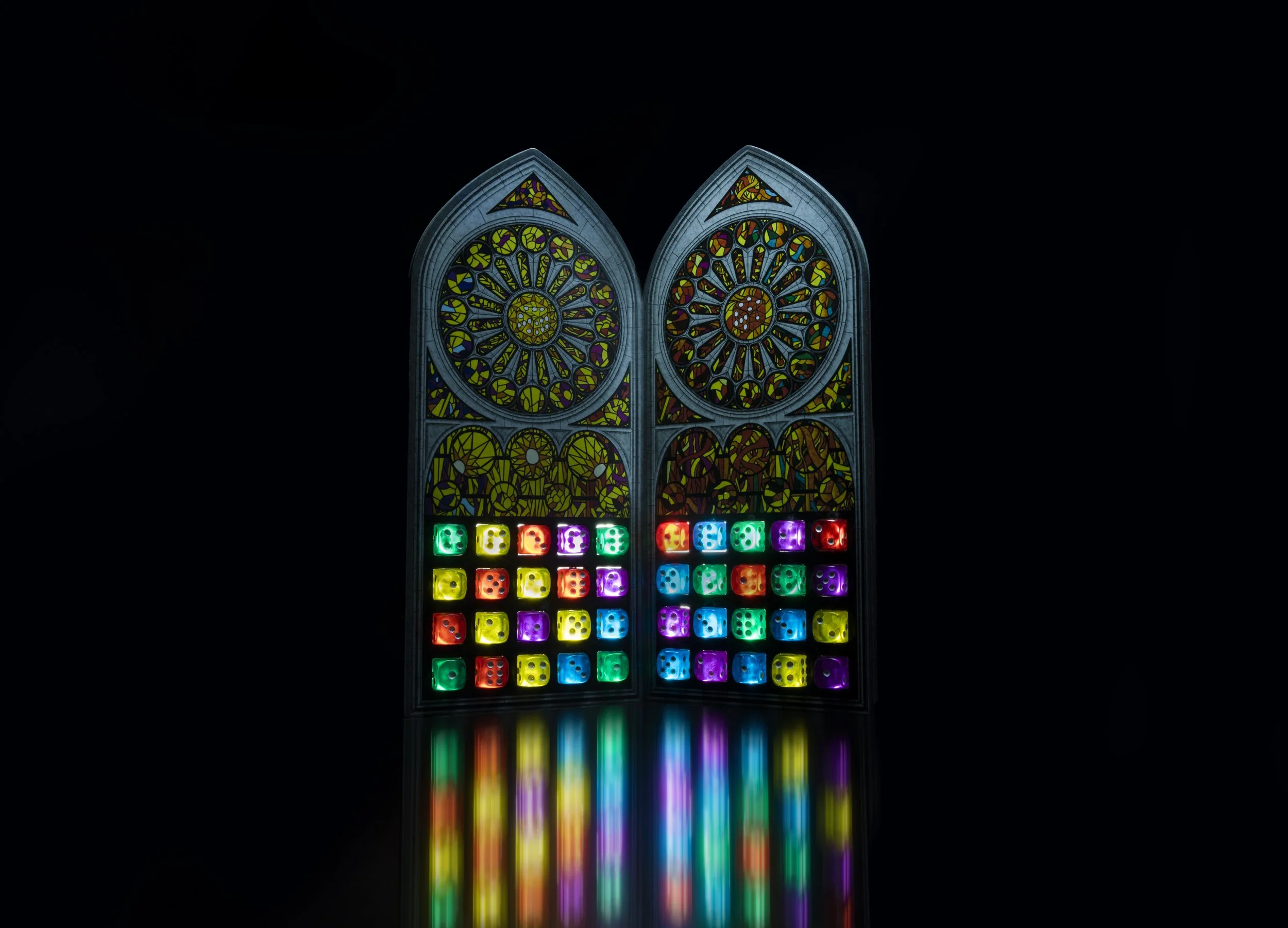 Colorful stained glass window with two circular rose windows at the top and illuminated dice below, casting a rainbow reflection on a dark surface.