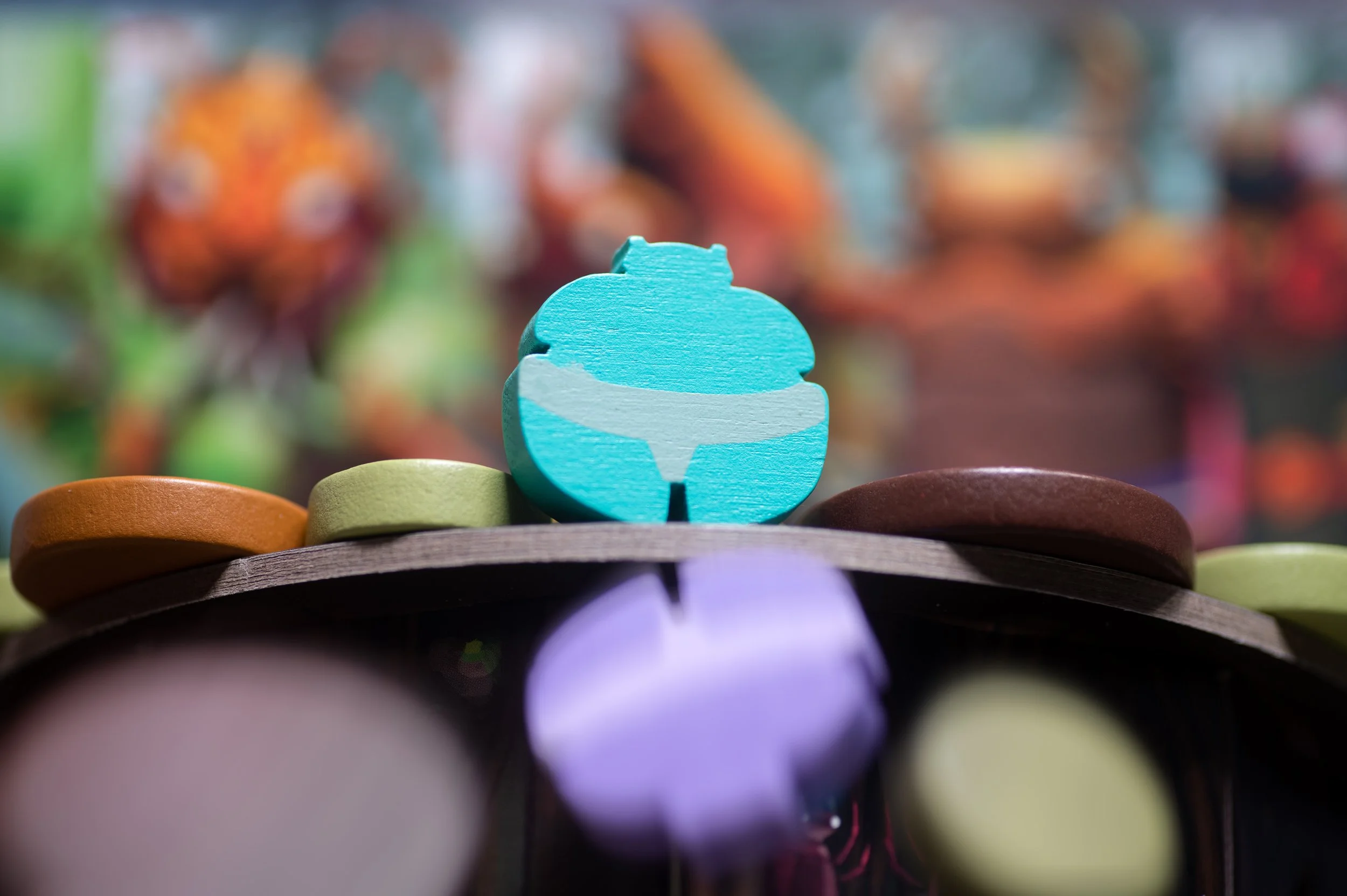 Close-up of a blue wooden game piece with a fish shape, surrounded by various colorful circular game pieces on a wooden surface with a blurred background of brightly colored fish.