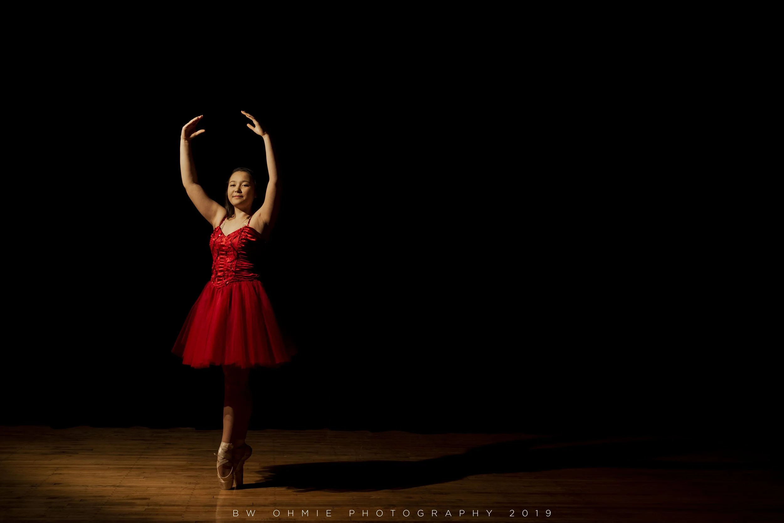 A ballerina in a red tutu performs on stage with a spotlight, standing on her toes with arms raised in a graceful pose.