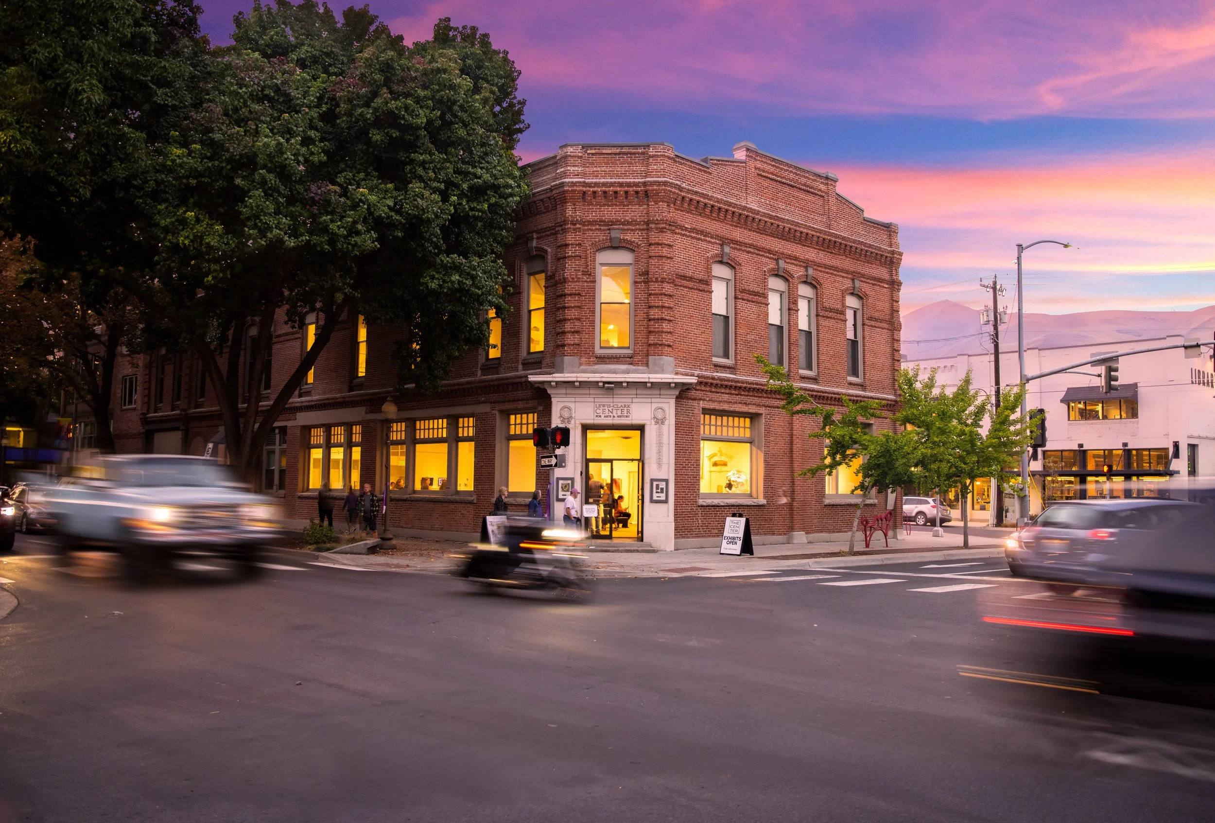 A corner building with a brick exterior and large illuminated windows at sunset, with cars and motorcycles passing by and a purple and pink sky in the background.