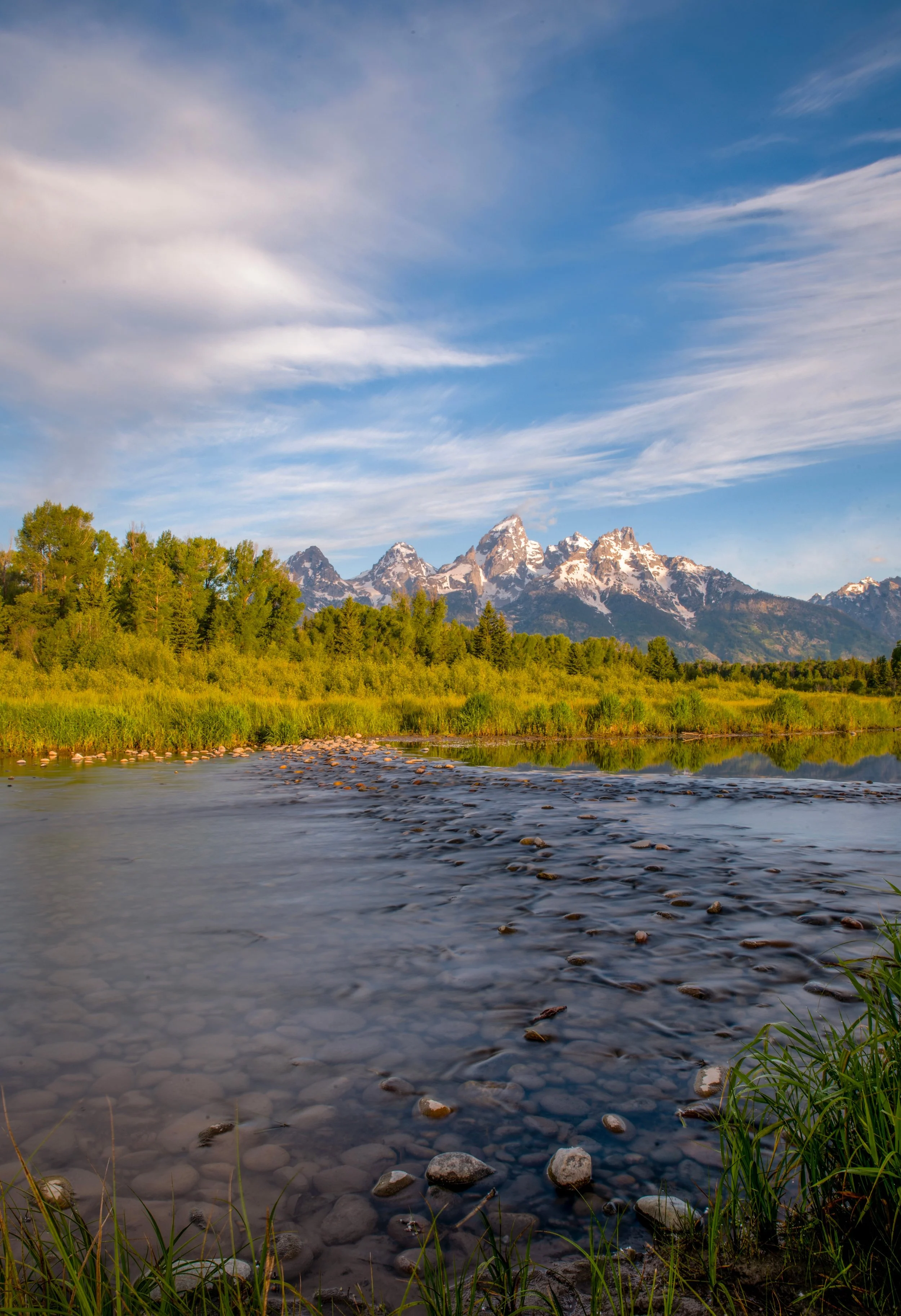 Mountain range with snow-capped peaks, green forest, river with rocks, and a blue sky with clouds.