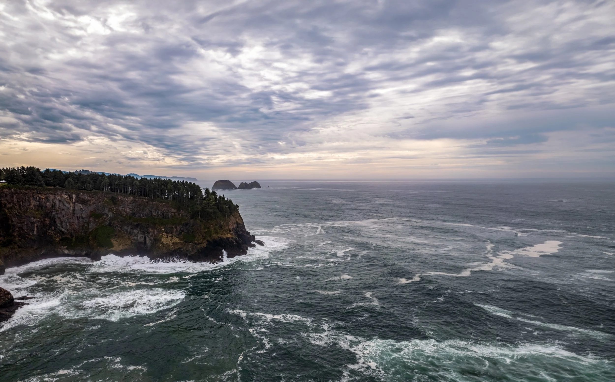 A rugged coastline with cliffs and trees, overlooking the ocean, under a partly cloudy sky with multiple layers of clouds and distant rock formations.