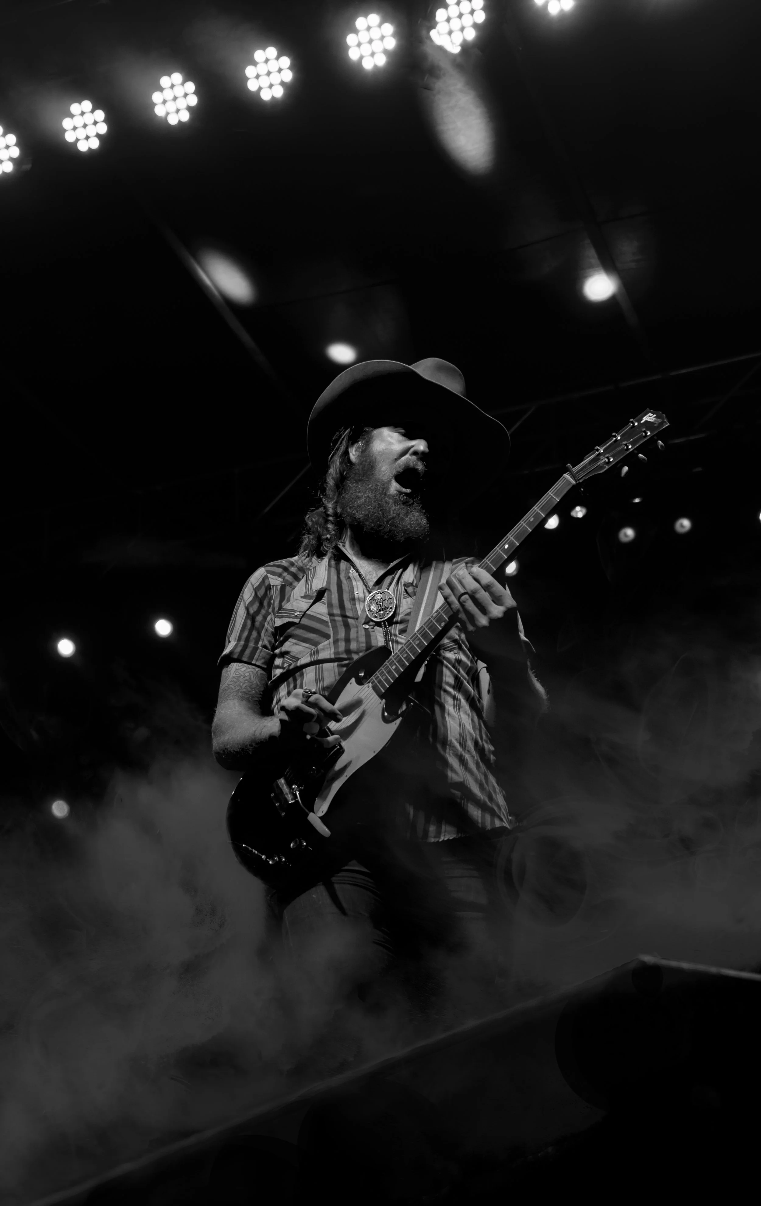 A musician with a beard, wearing a cowboy hat, striped shirt, and bolo tie, plays an electric guitar on stage with stage lights overhead.