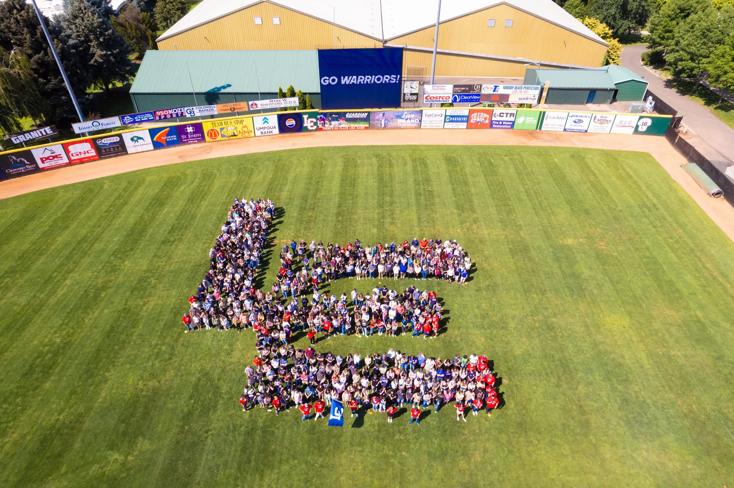 A large group of people gathered on a baseball field. The field is green with a dirt warning track, and there are advertisements and a large banner reading 'GO WARRIORS!' along the outfield fence. Buildings and trees are visible in the background.
