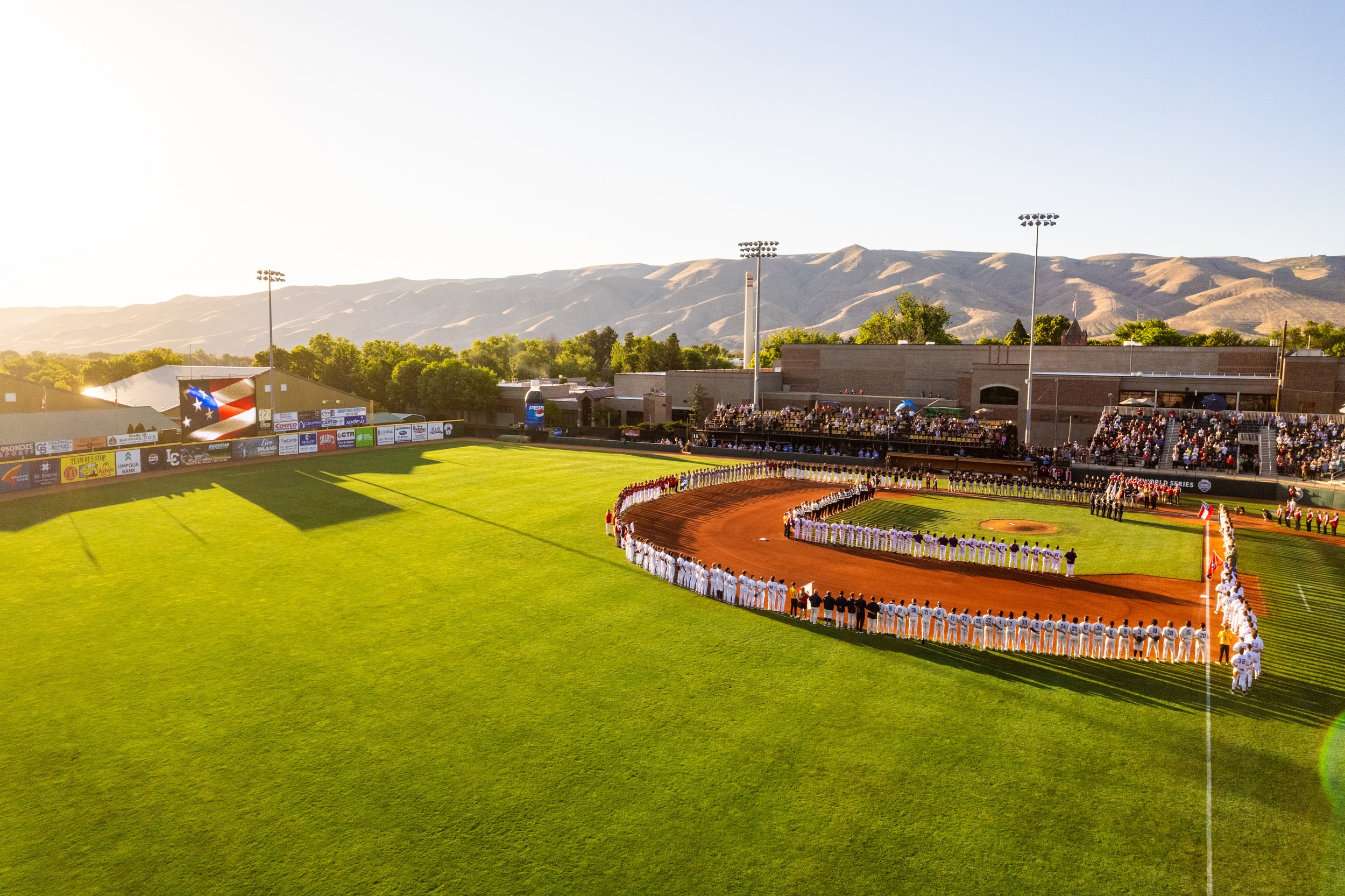 A baseball game at a stadium during sunset, with players standing in line on the field and the audience in the stands, banners along the outfield, and mountains in the background.