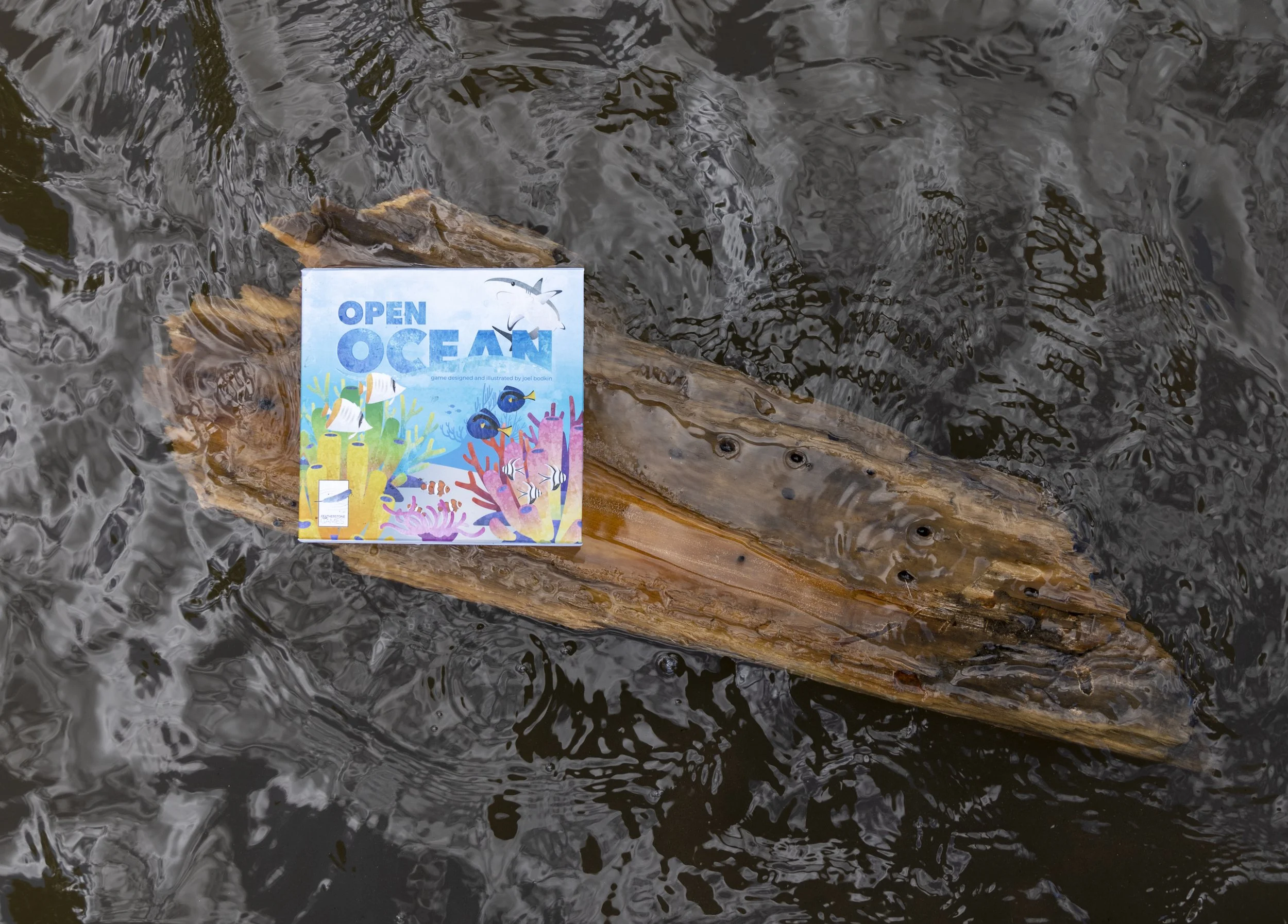 A small wooden boat floating in dark water with a colorful game box titled 'Open Ocean' placed on top of it.