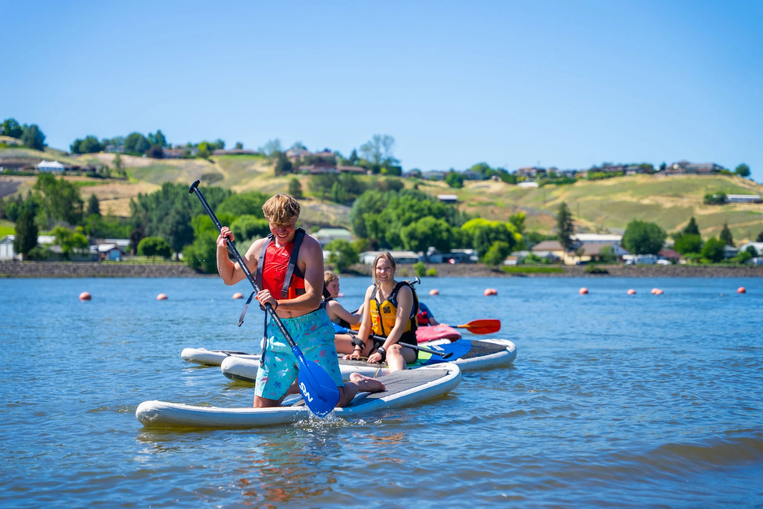 Four people, two women and two children, on paddleboards in a lake with hills and houses in the background.