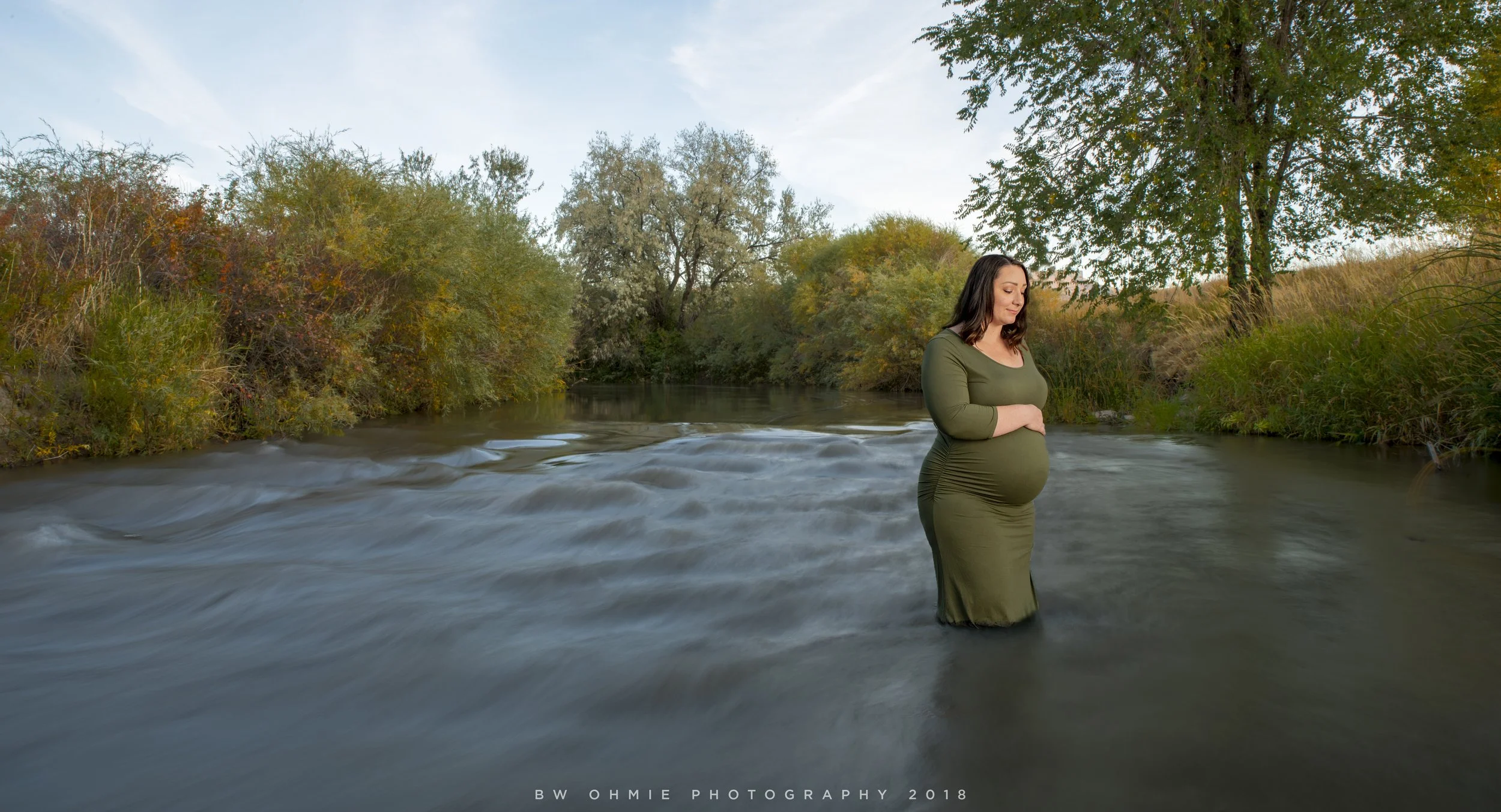 Pregnant woman standing in a river with her eyes closed and hands resting on her belly, surrounded by trees with autumn foliage.