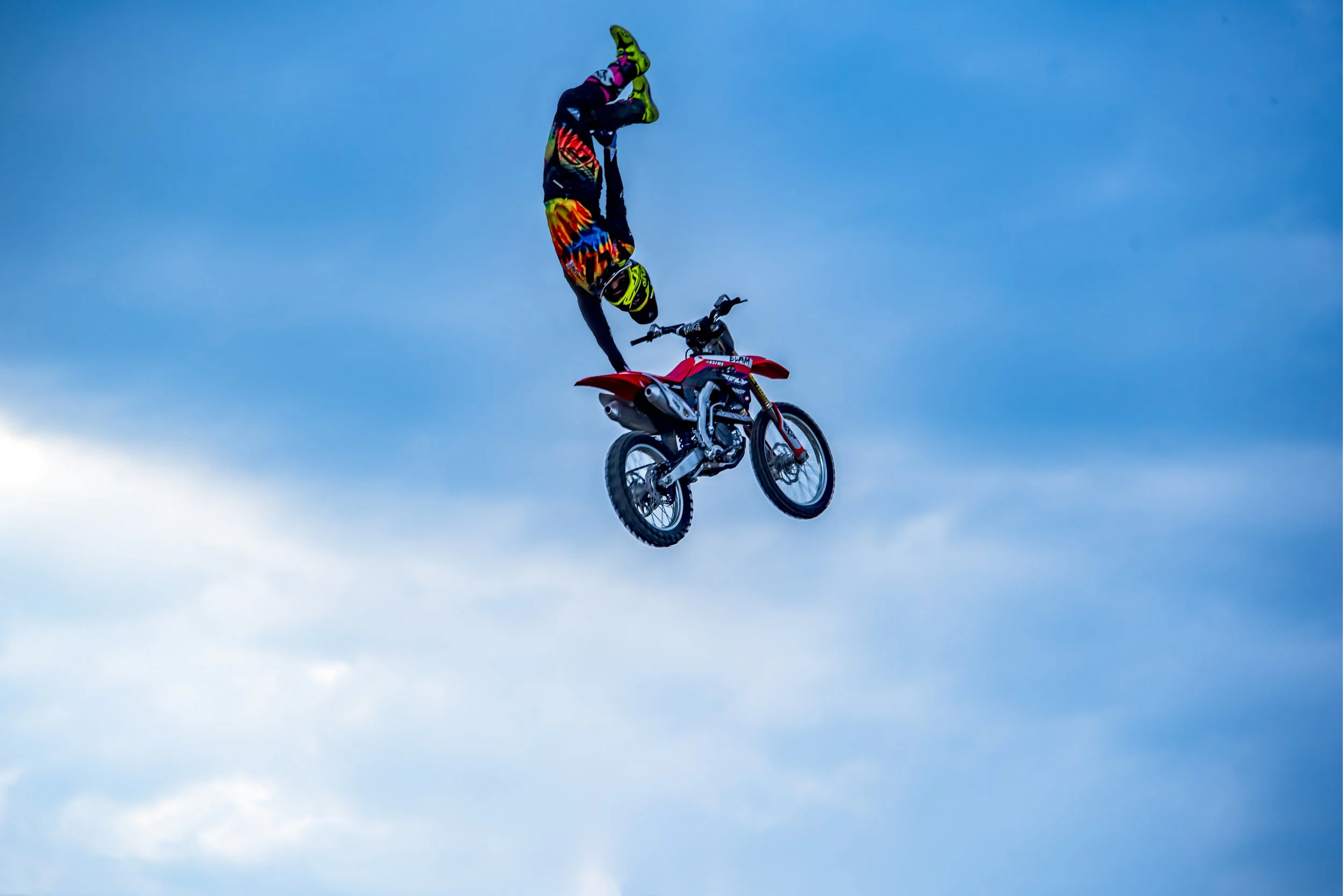 A motocross rider performing a mid-air stunt on a red dirt bike against a blue sky with clouds.