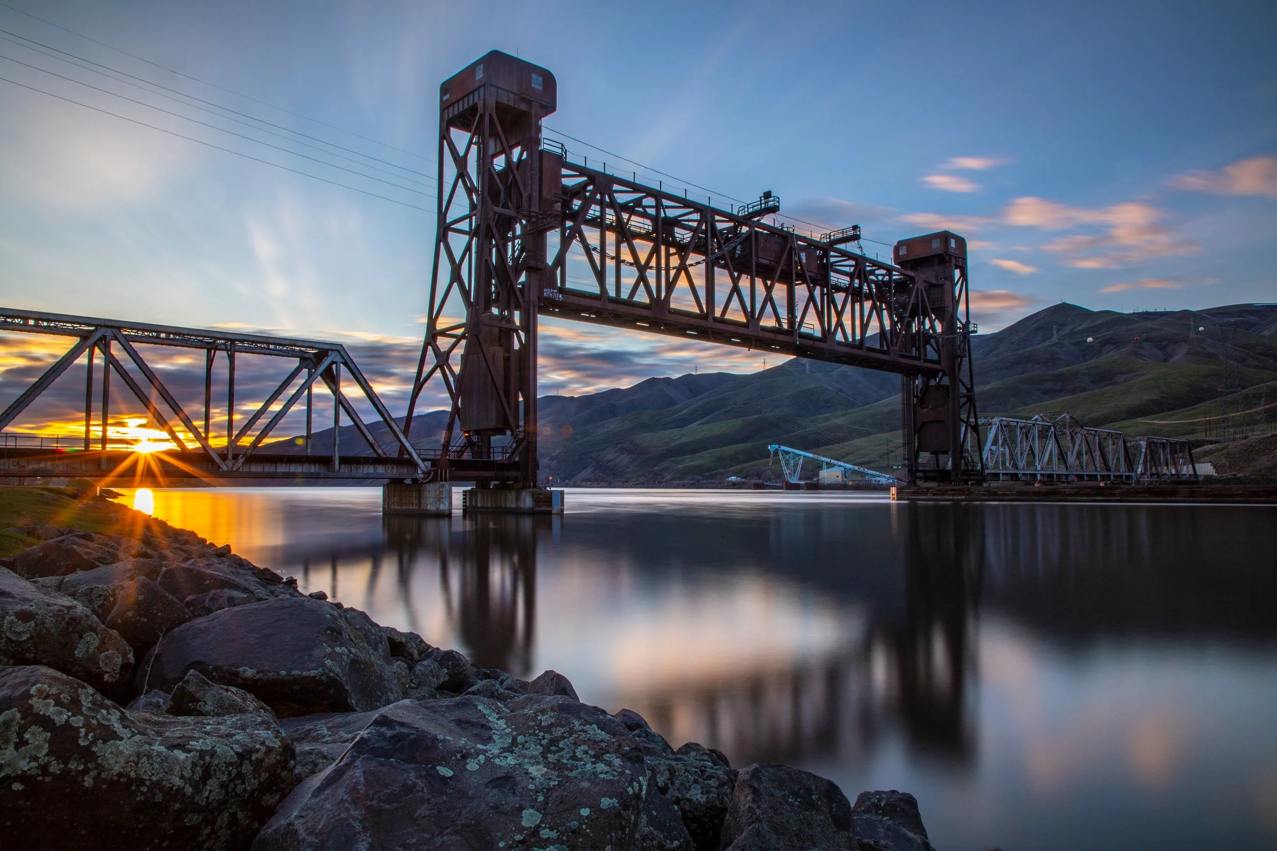 Steel lift bridge over water at sunset with mountains in the background.