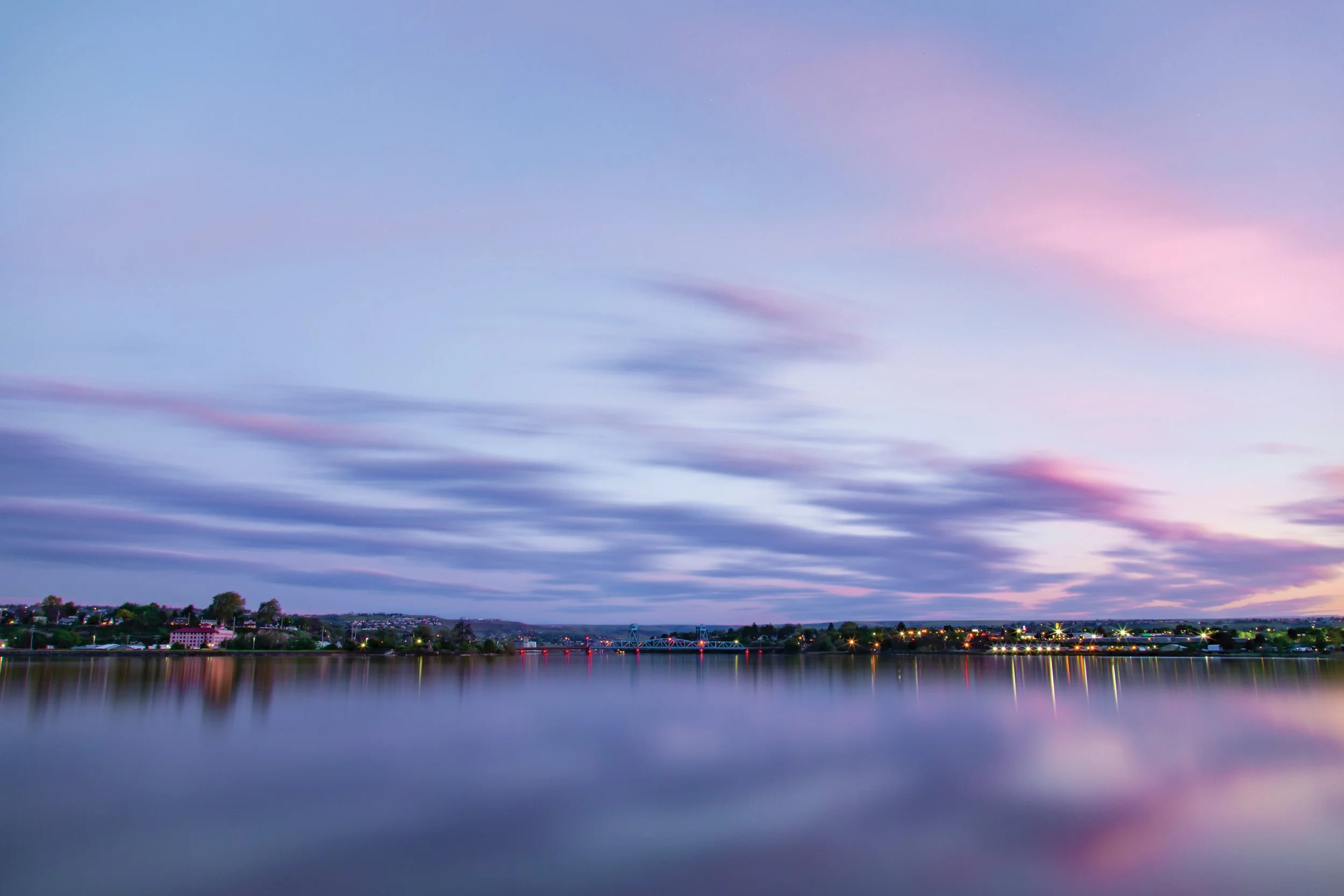 Calm river with city lights and buildings in the distance during sunset, with pink and blue sky and blurred clouds.