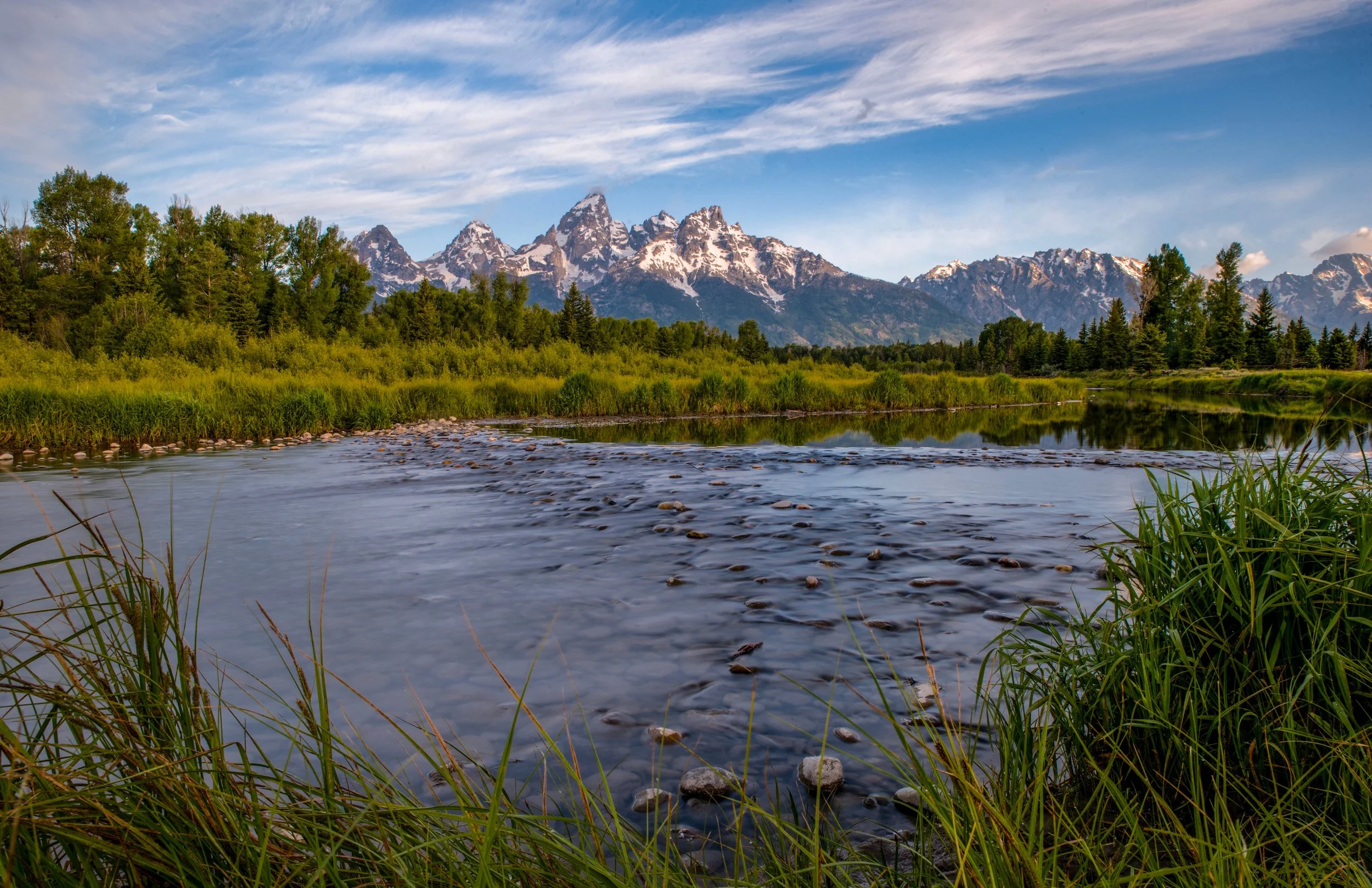 Scenic landscape of the Grand Tetons with a river in the foreground, green trees, and snow-capped mountains in the background under a partly cloudy sky.