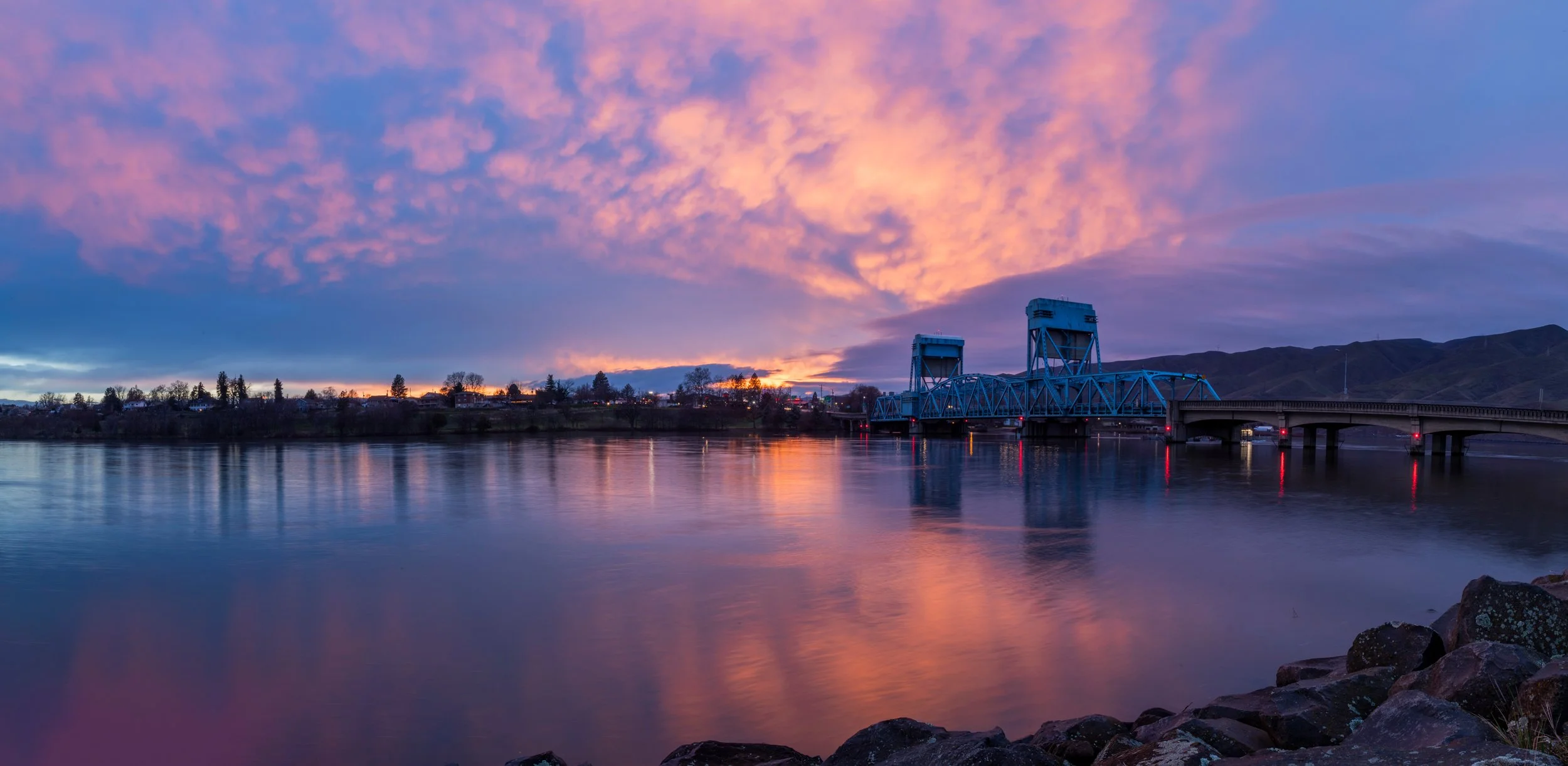 Sunset over a river with a blue drawbridge and mountains in the background.