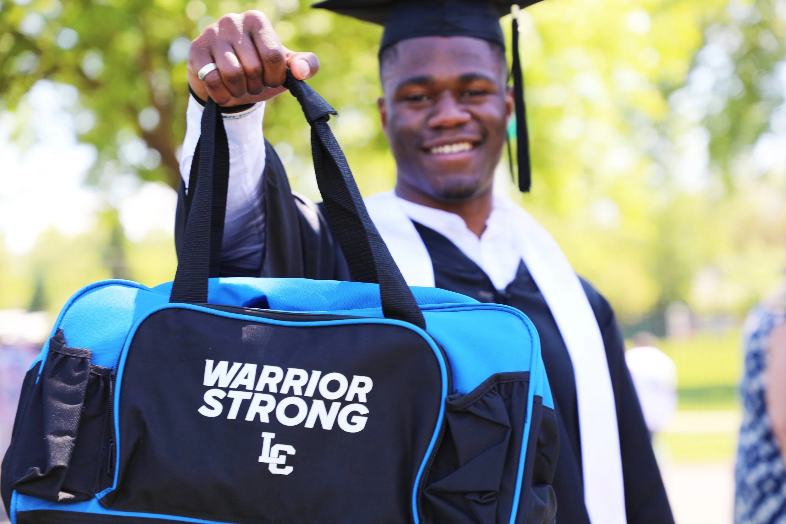 A young man in a graduation cap and gown holding a blue sports bag with the words 'Warrior Strong' and the initials 'LC' on it, outdoors with trees in the background.
