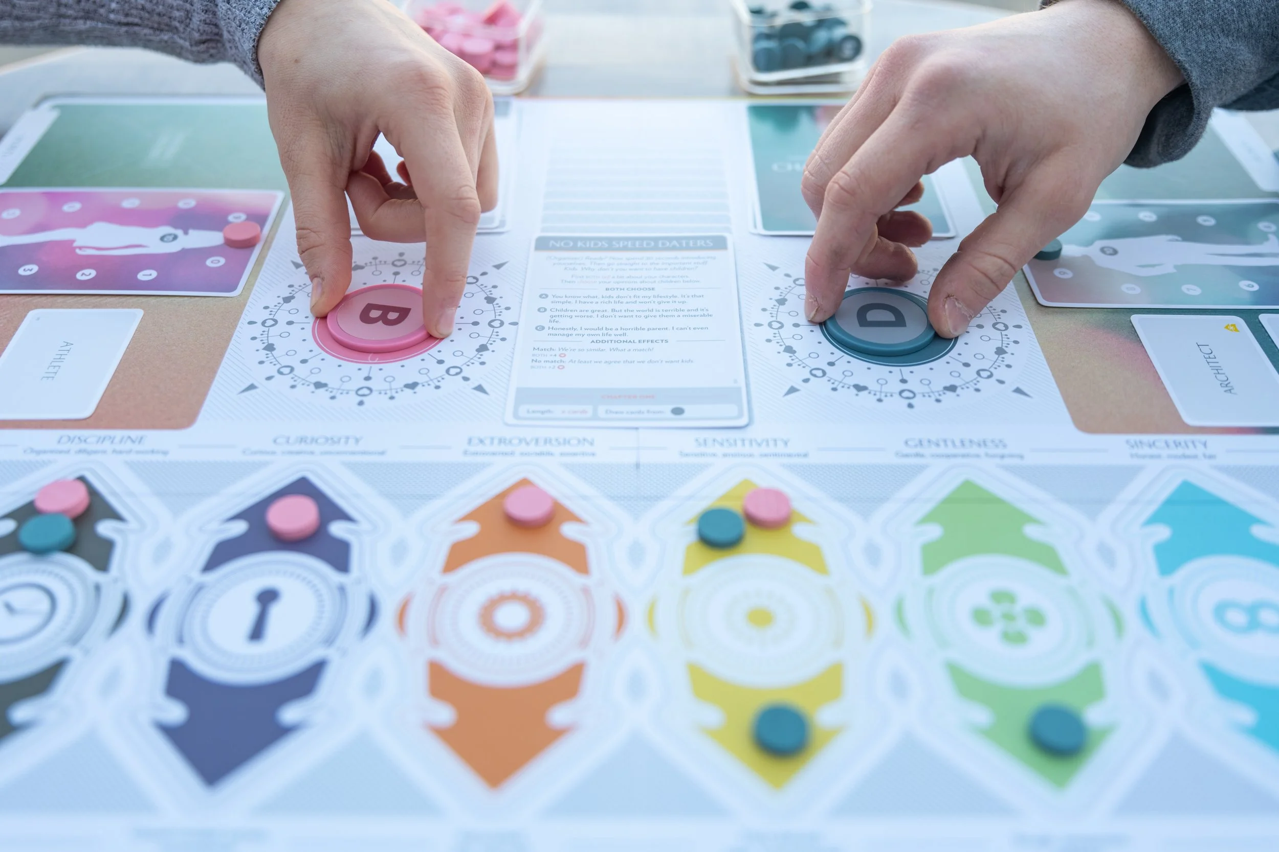 Close-up of two hands playing a tabletop board game called 'Fog of Love'. The game features colorful discs marked with letters 'B' and 'D' and sequence cards with colorful patterns. The game surface has various personality traits written across it.
