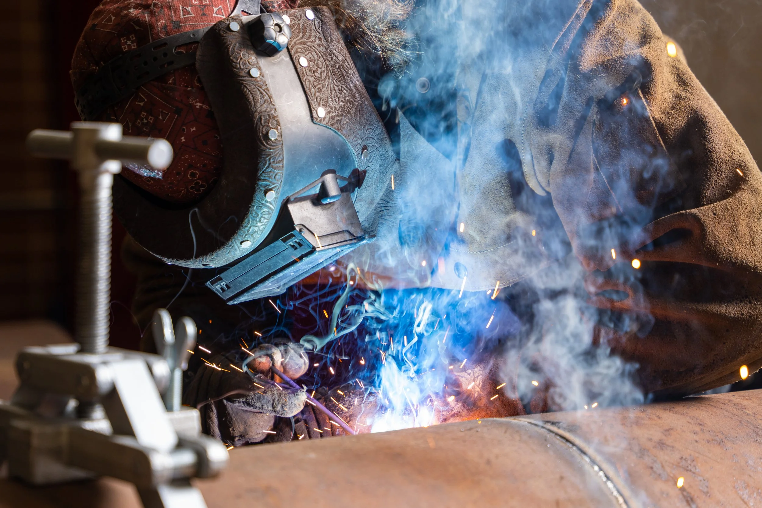 A person welding a metal pipe, wearing protective gloves, a welding helmet that is lifted, and a brown jacket, with sparks and blue light from the welding process visible.