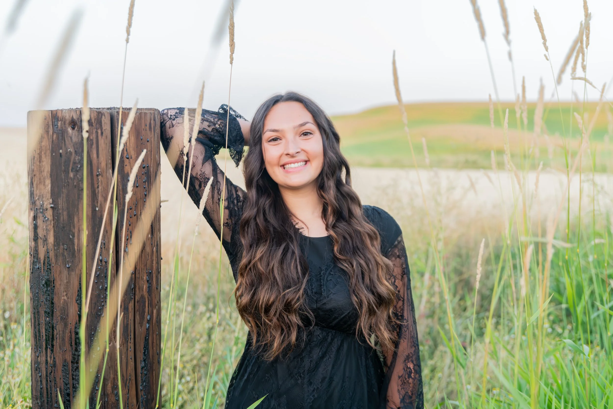 Young woman in a black lace dress smiling outdoors in a grassy field with rolling hills in the background.