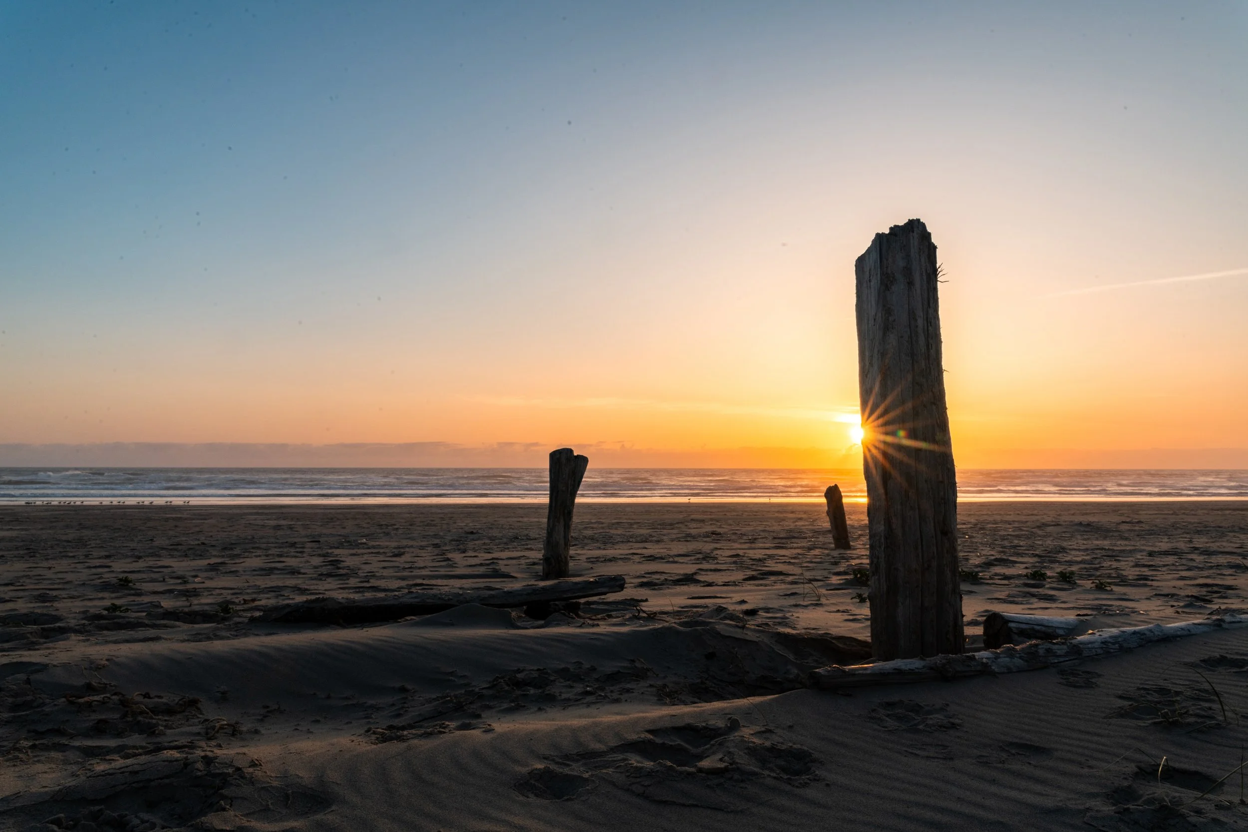Sunset on a beach with weathered wooden posts sticking out of the sand and a calm ocean in the background.