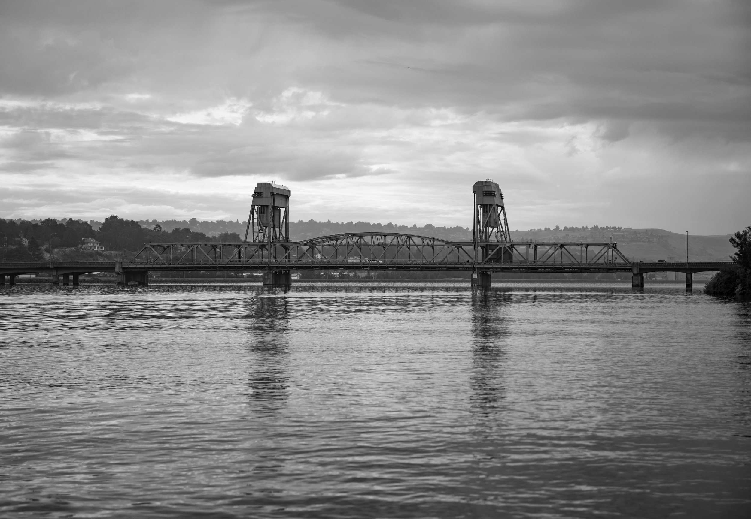 Black and white photo of a suspension bridge crossing a body of water with hills in the background and a cloudy sky.