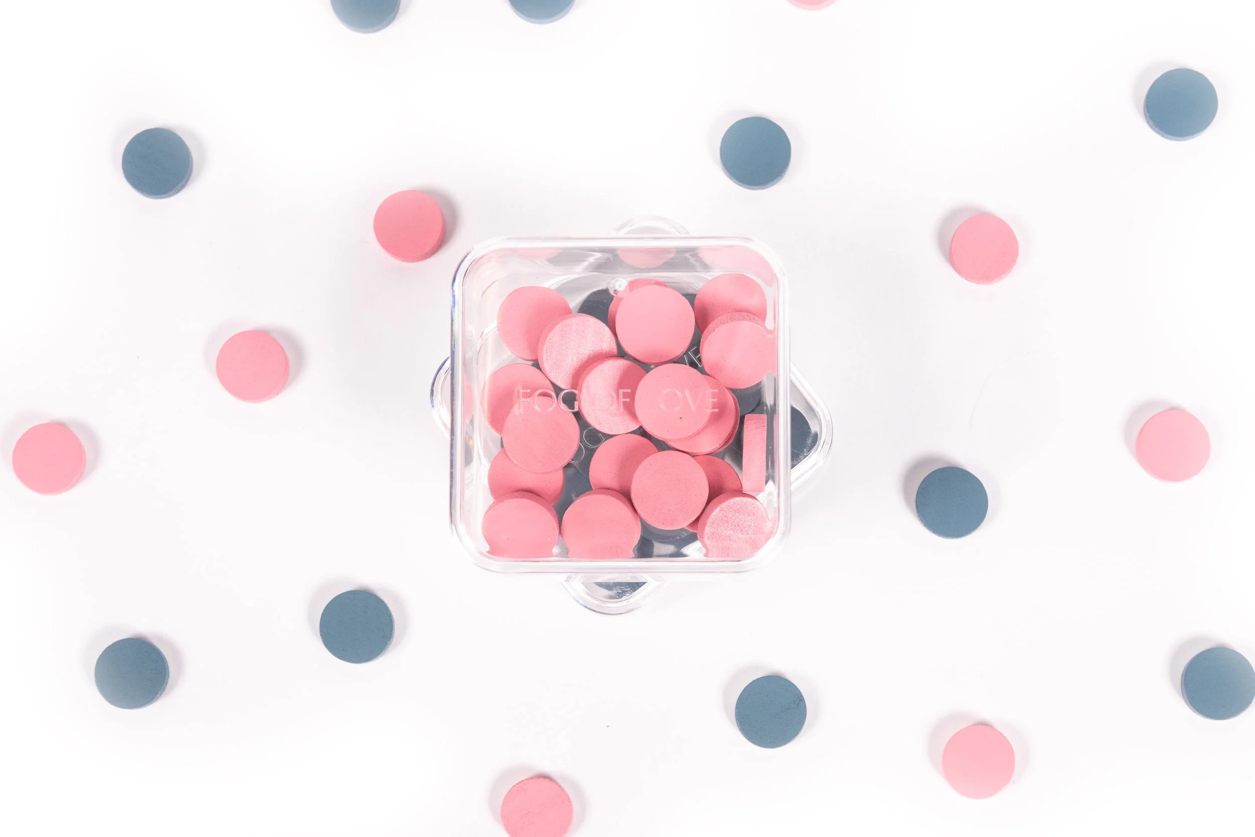 A clear plastic container filled with pink circular candies or tablets, surrounded by pink and blue paper confetti on a white surface.