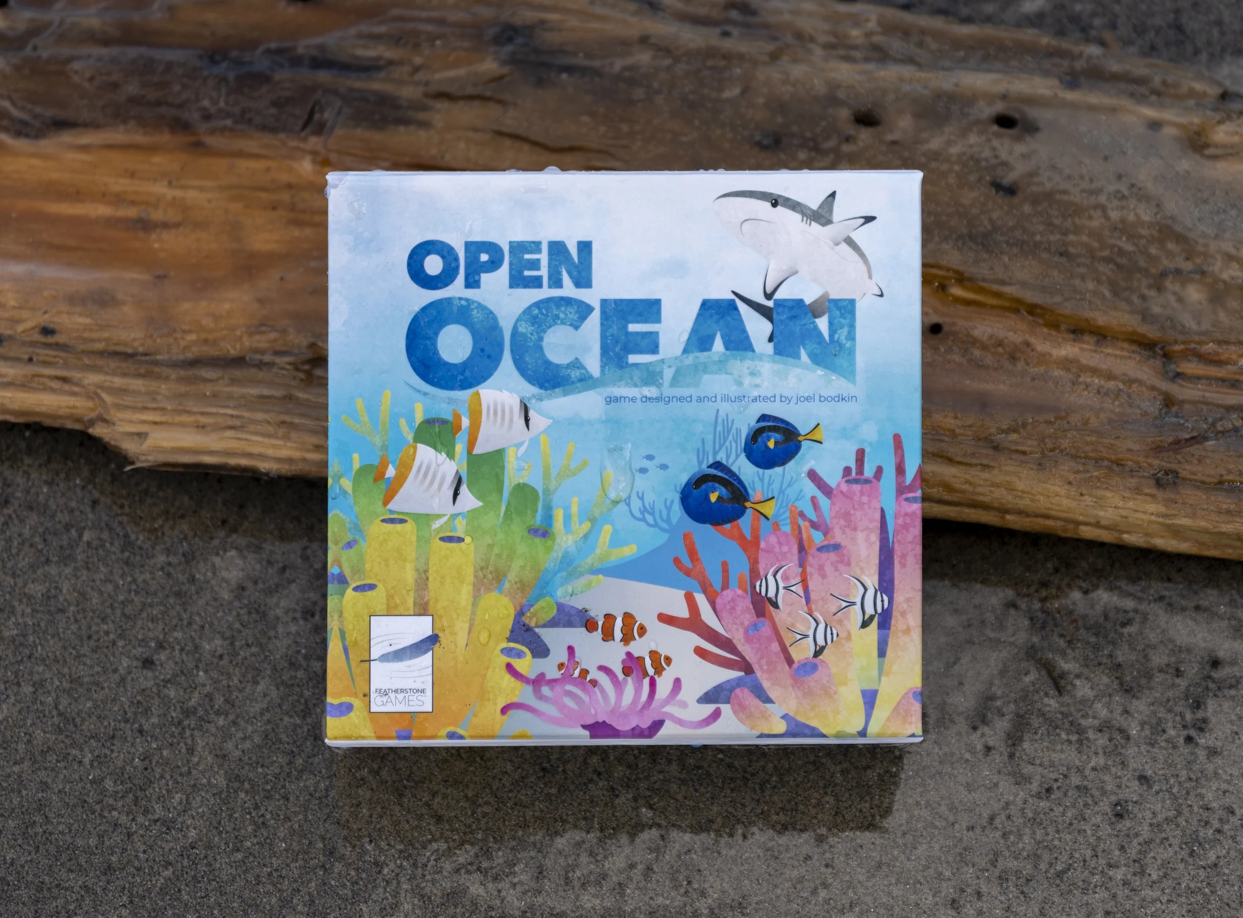 Box of a card game titled 'Open Ocean' with colorful underwater fish and coral illustrations, placed on sand with a piece of driftwood in the background.