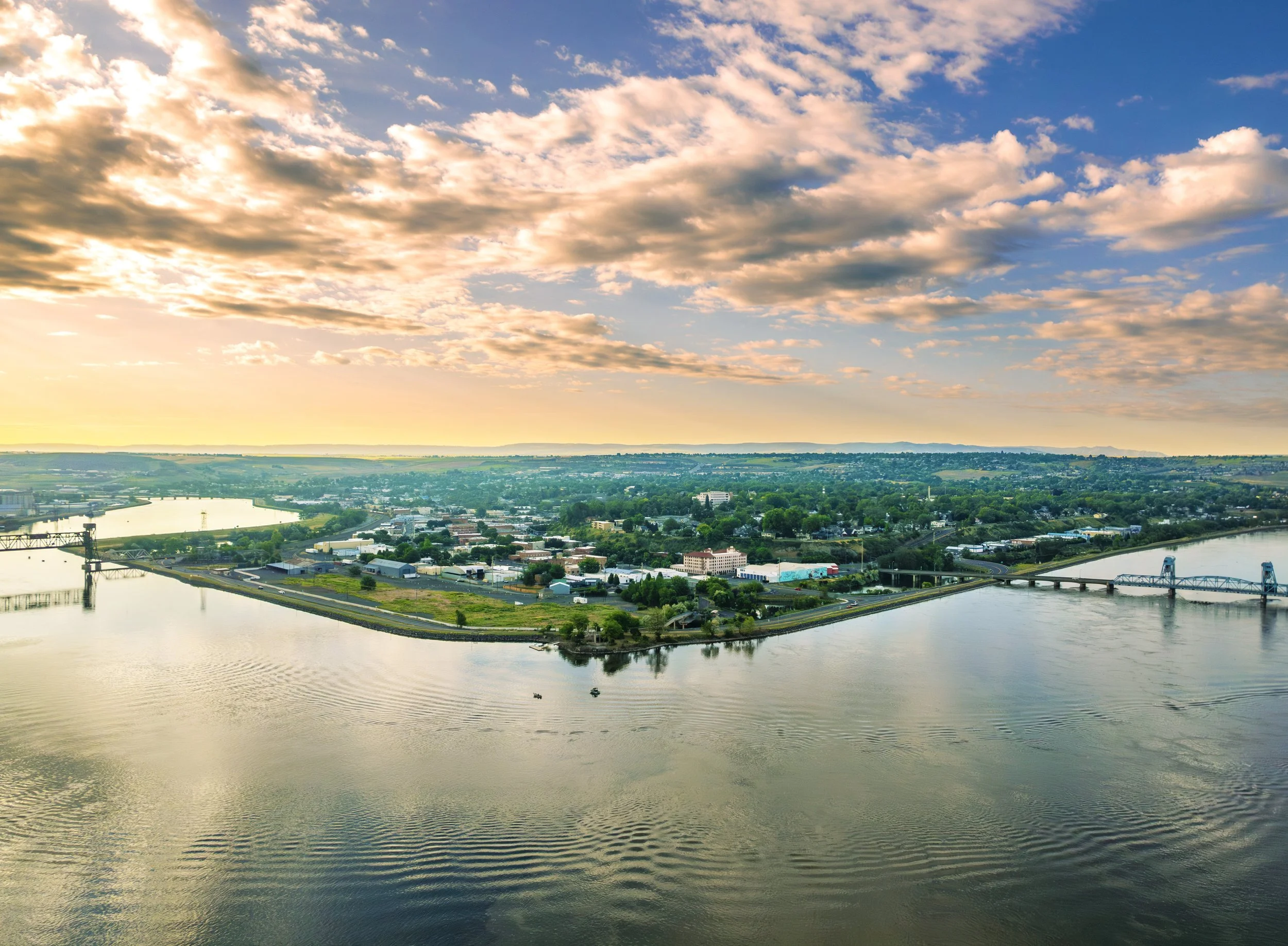 Aerial view of a city with a river, bridges, and green areas during sunset, with clouds in the sky.