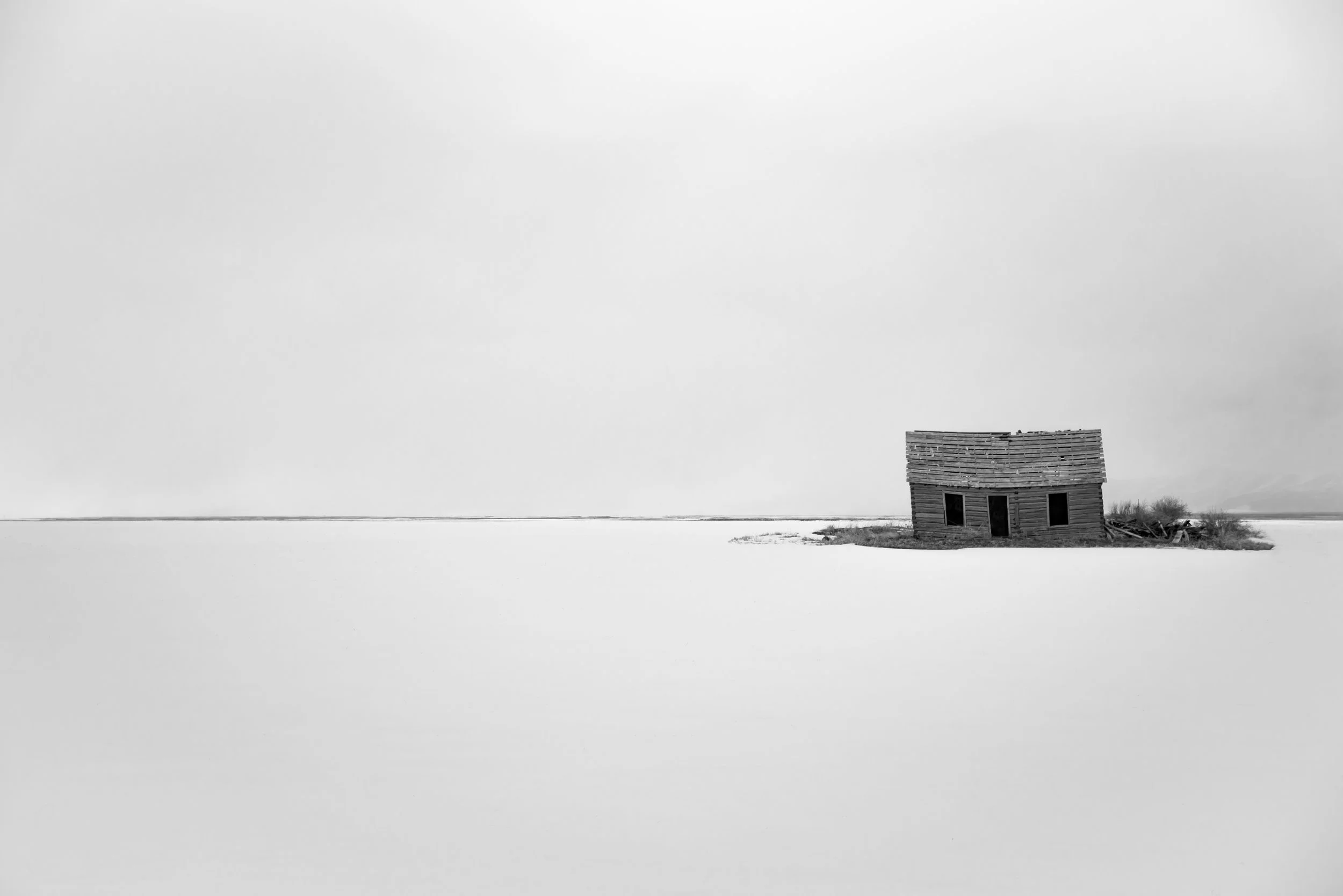 A solitary wooden house tilted and dilapidated in a vast, snow-covered landscape under a cloudy sky.